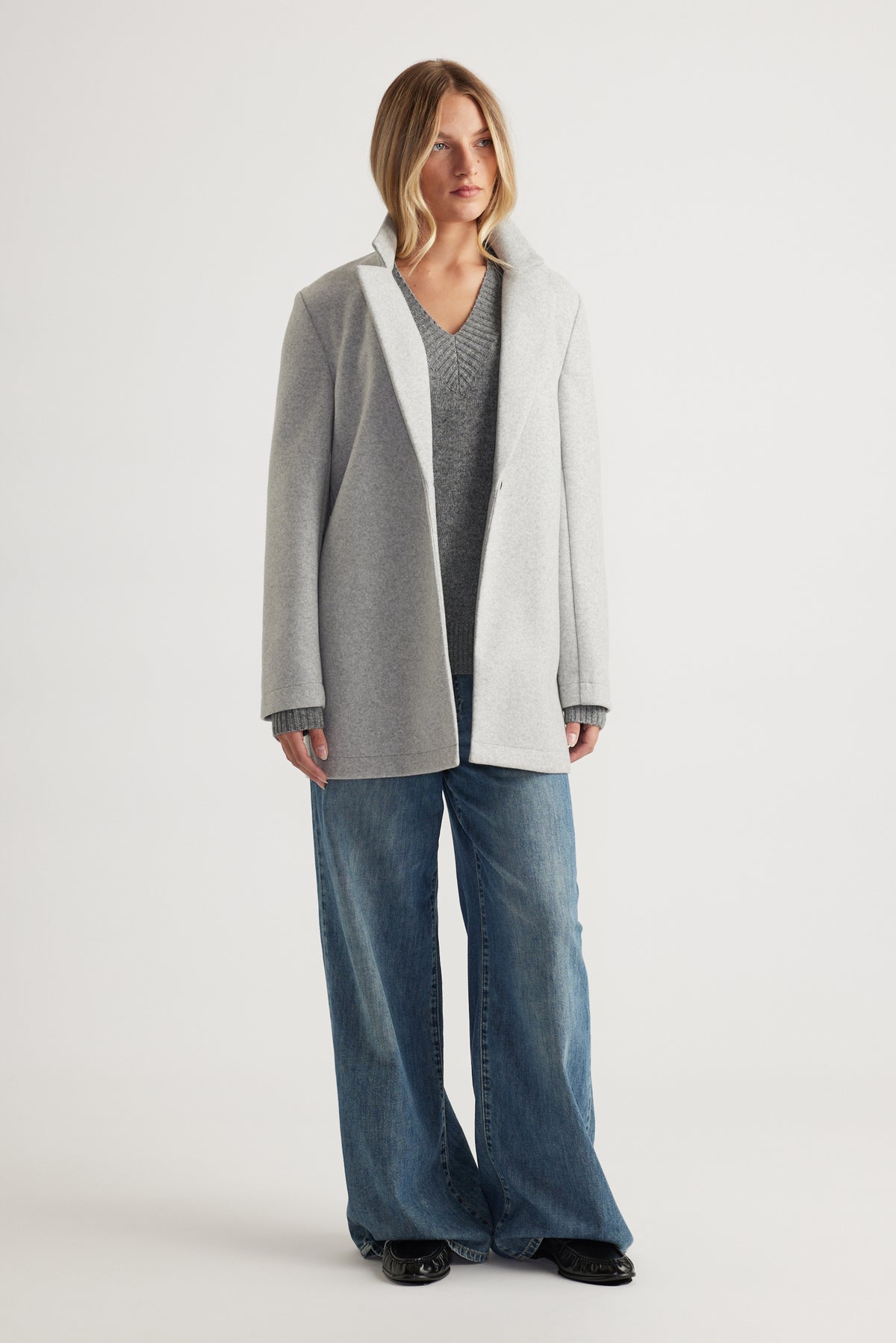 Woman wearing a light grey mid-length coat layered over a grey knit sweater and wide-leg blue denim jeans for a casual look