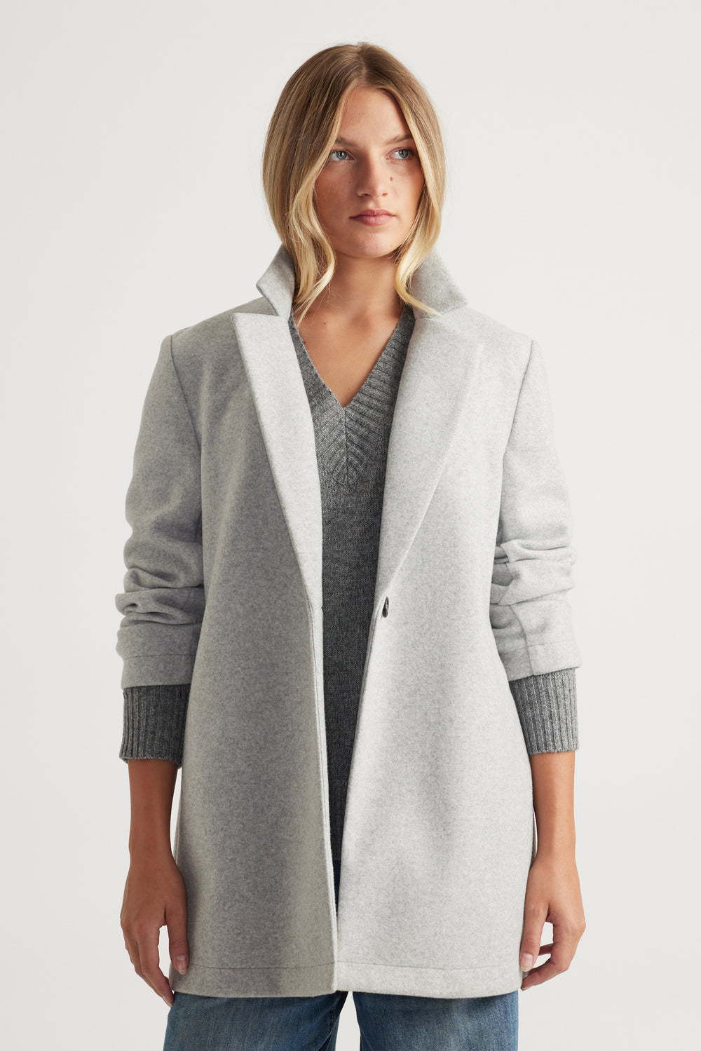 Woman wearing a light grey mid-length felt coat with a wide collar over a textured grey knit top and blue jeans