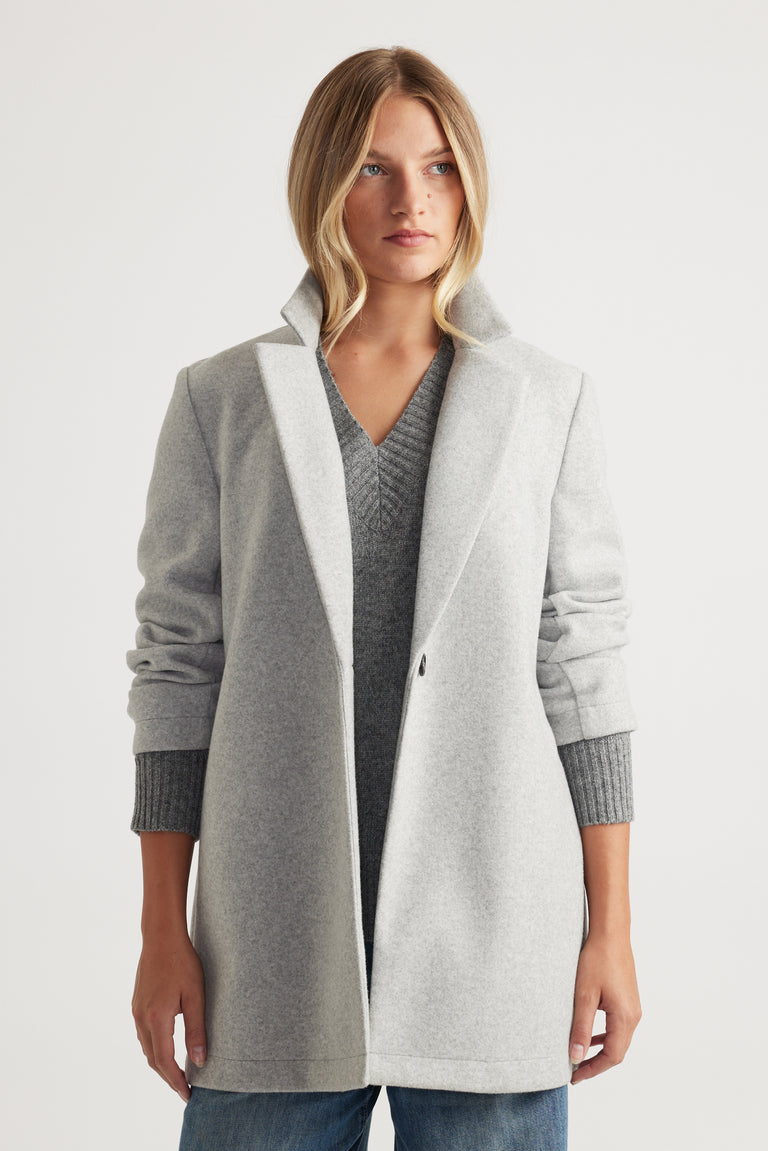 Woman wearing a light grey mid-length felt coat with a wide collar over a textured grey knit top and blue jeans