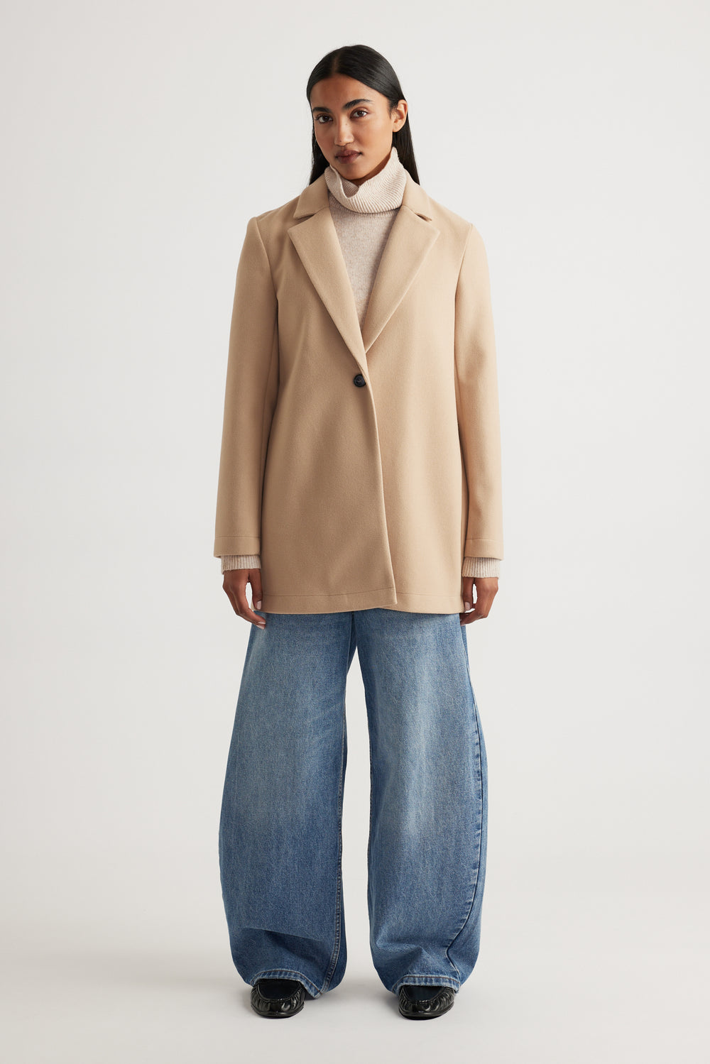 Woman wearing a soft camel mid length coat layered over a beige turtleneck paired with wide-leg denim jeans for a modern look