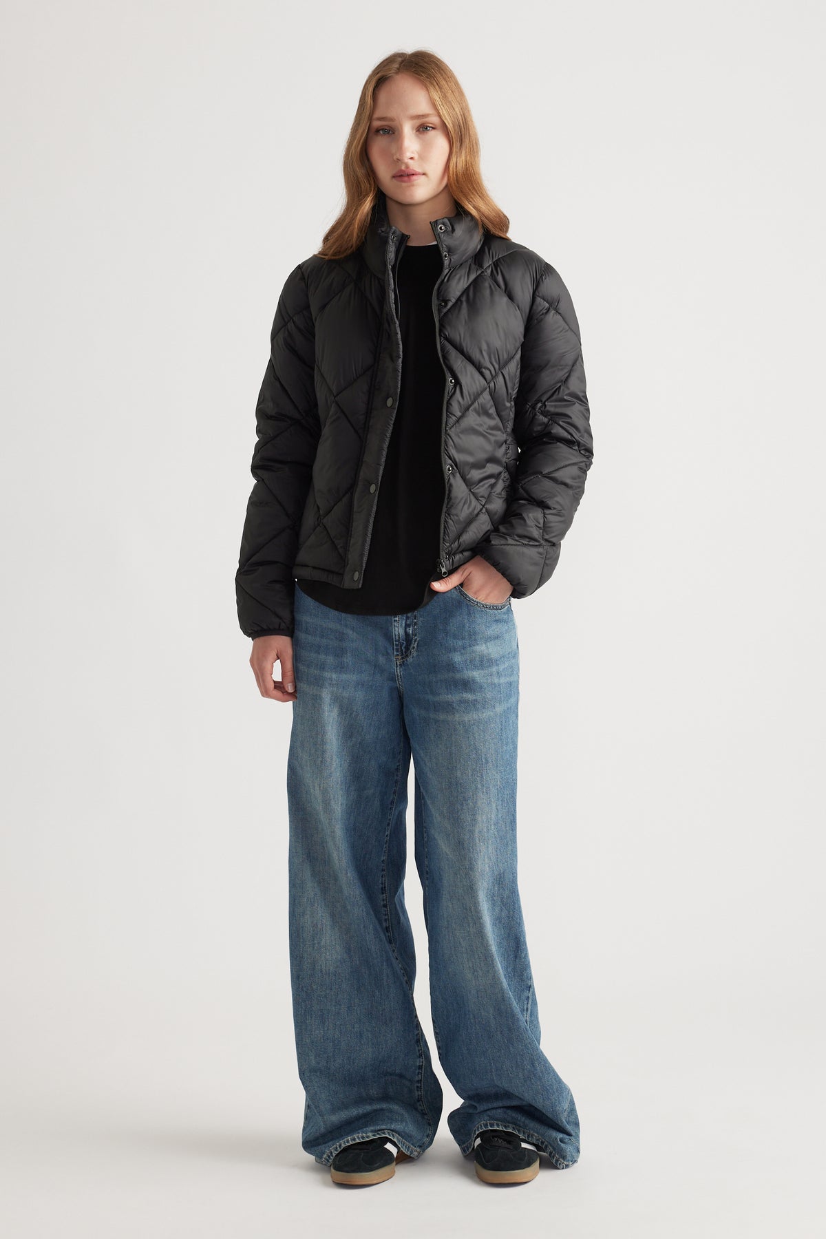 Woman wearing a black short puffer jacket layered over a black top, paired with wide-leg blue denim jeans and black sneakers.