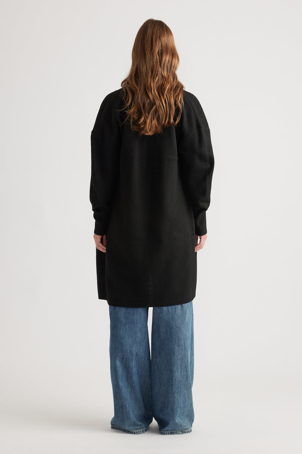 Woman showcasing a loose black longline cardigan paired with wide-leg blue denim jeans in a relaxed style from the back view.