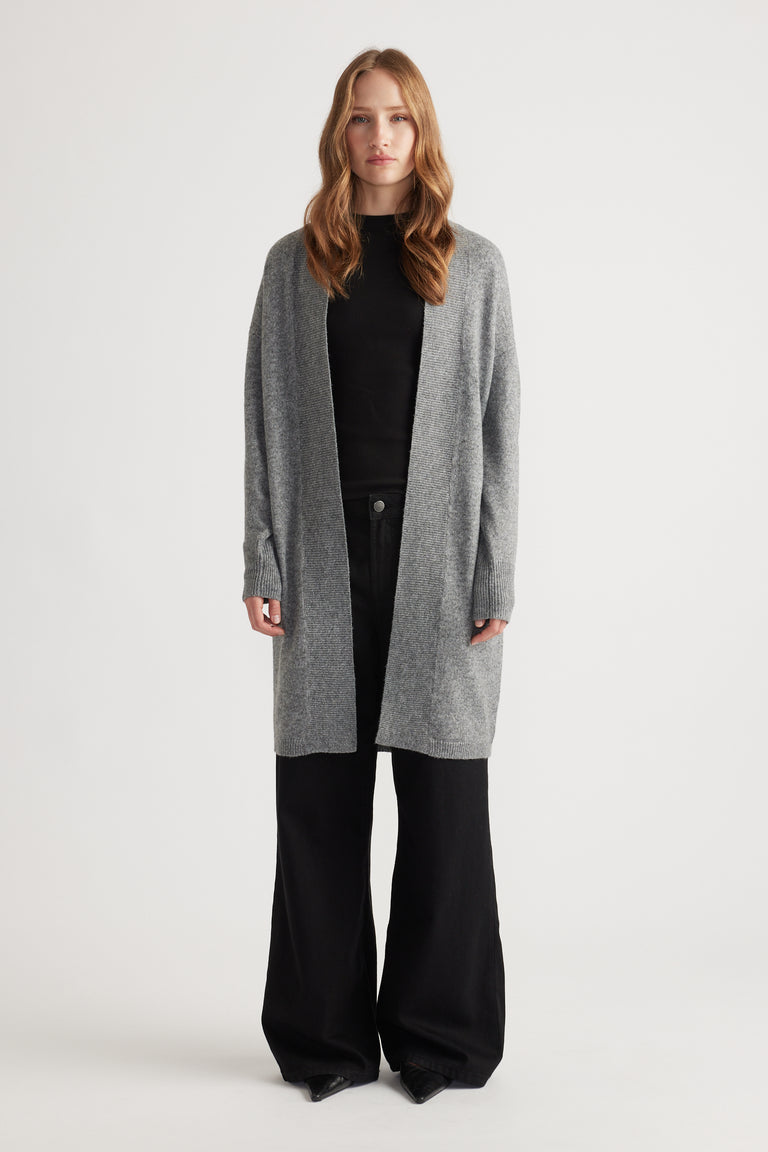 Woman wearing mid-grey longline knit cardigan over black top and wide-leg black pants against white background