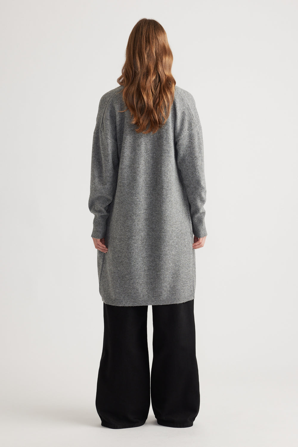 Woman shown from behind wearing a mid-grey longline knit cardigan paired with loose black pants against a plain backdrop