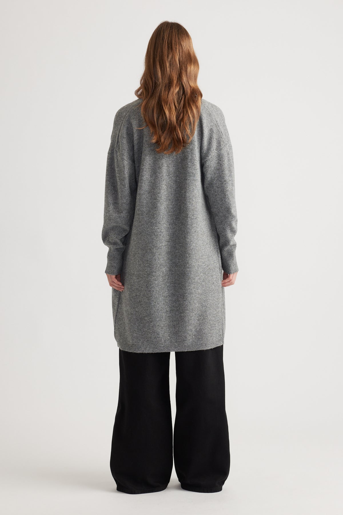 Woman shown from behind wearing a mid-grey longline knit cardigan paired with loose black pants against a plain backdrop
