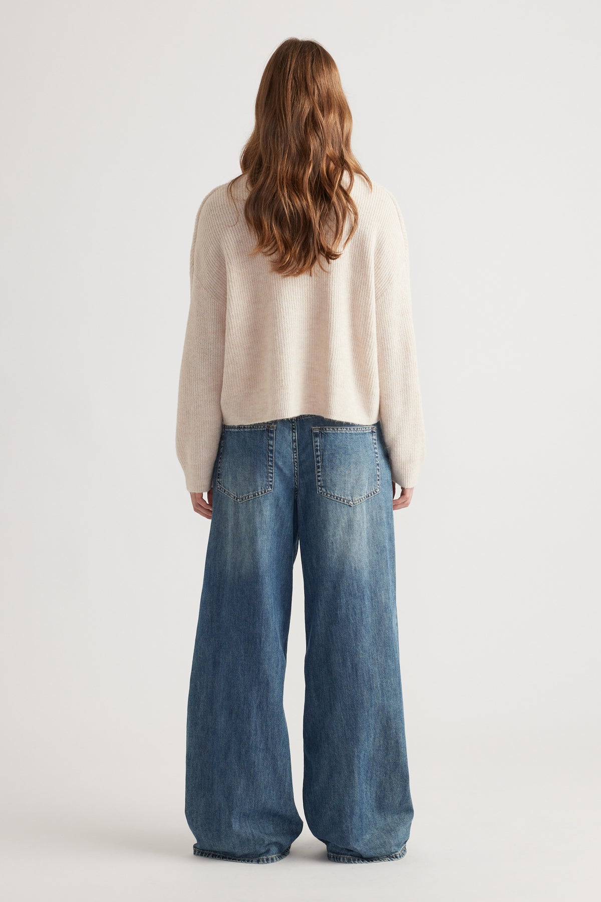 Woman with wavy hair wearing a cozy beige knit cardigan paired with relaxed wide-leg blue denim jeans from behind viewpoint