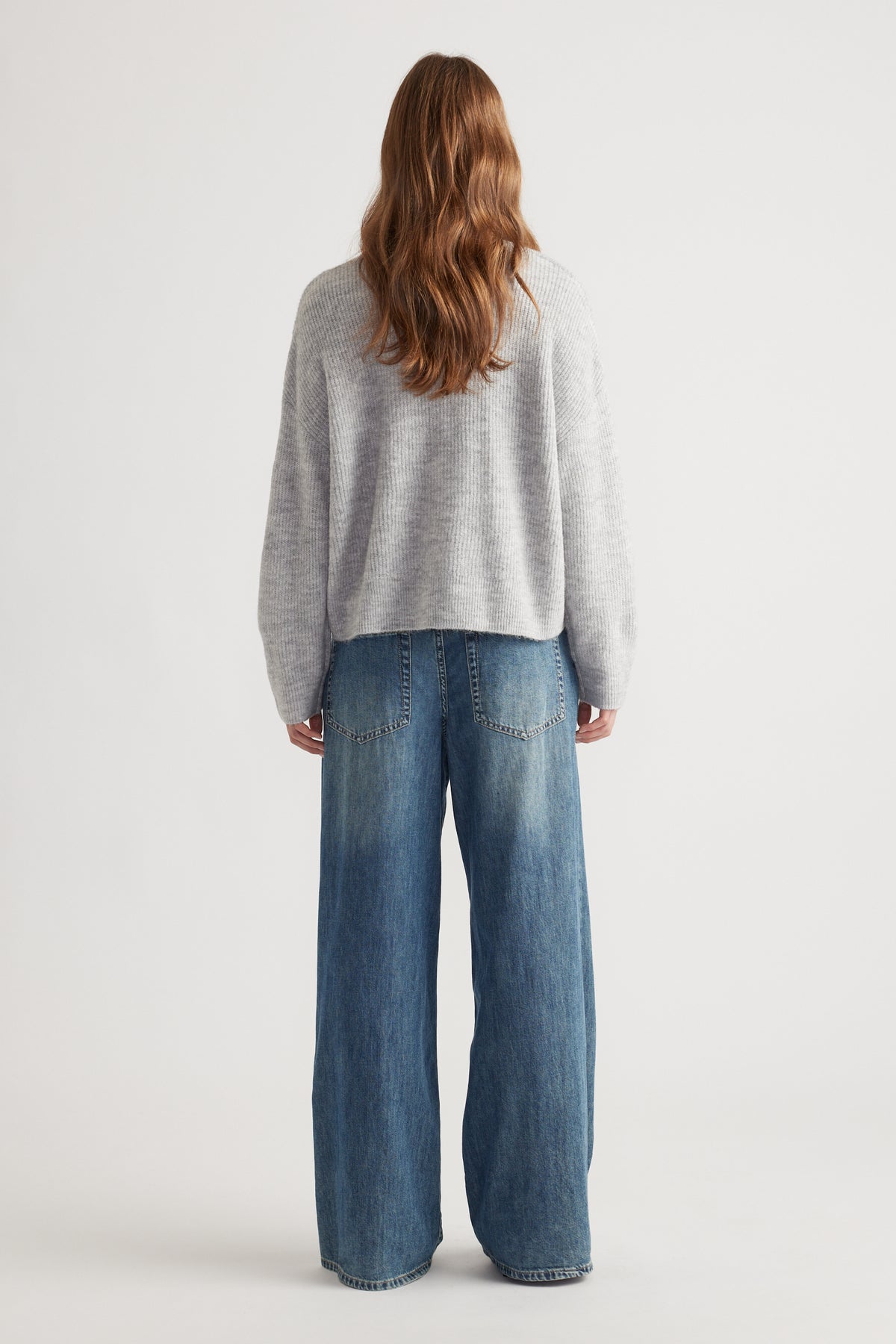 Woman with loose waves wearing a cozy light grey knit cardigan paired with wide-leg faded blue denim jeans viewed from behind