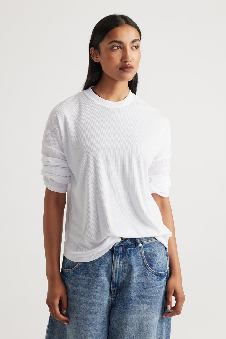 Woman wearing a relaxed white long-sleeve rib top paired with loose-fitting blue denim jeans against a plain background