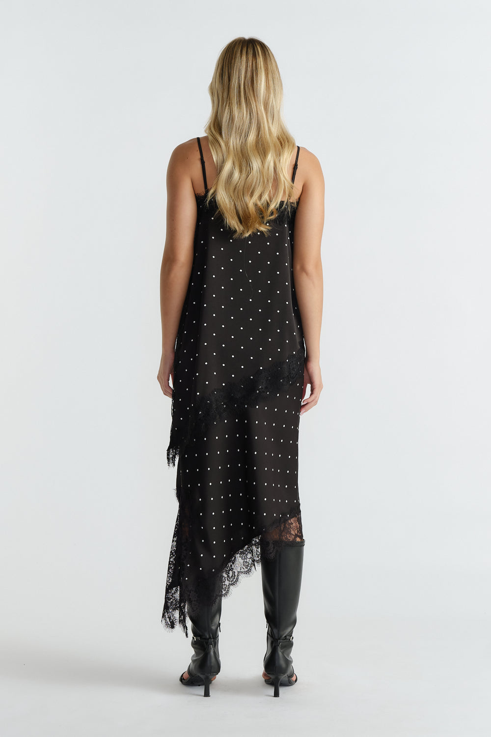 Woman wearing a black polka dot tunic with lace trim, thin straps, asymmetric hem, and black knee-high boots, back view