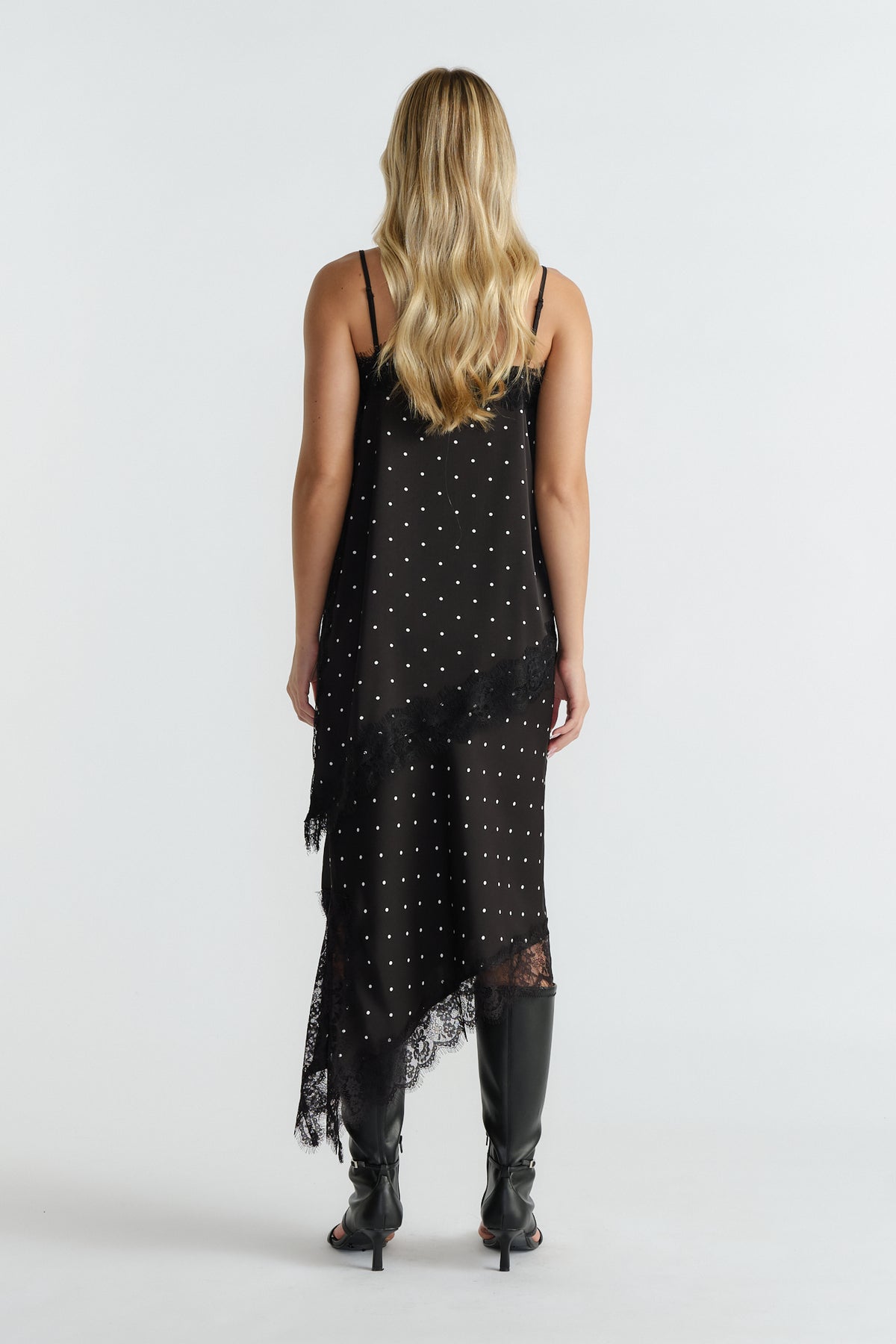 Woman wearing a black polka dot tunic with lace trim, thin straps, asymmetric hem, and black knee-high boots, back view