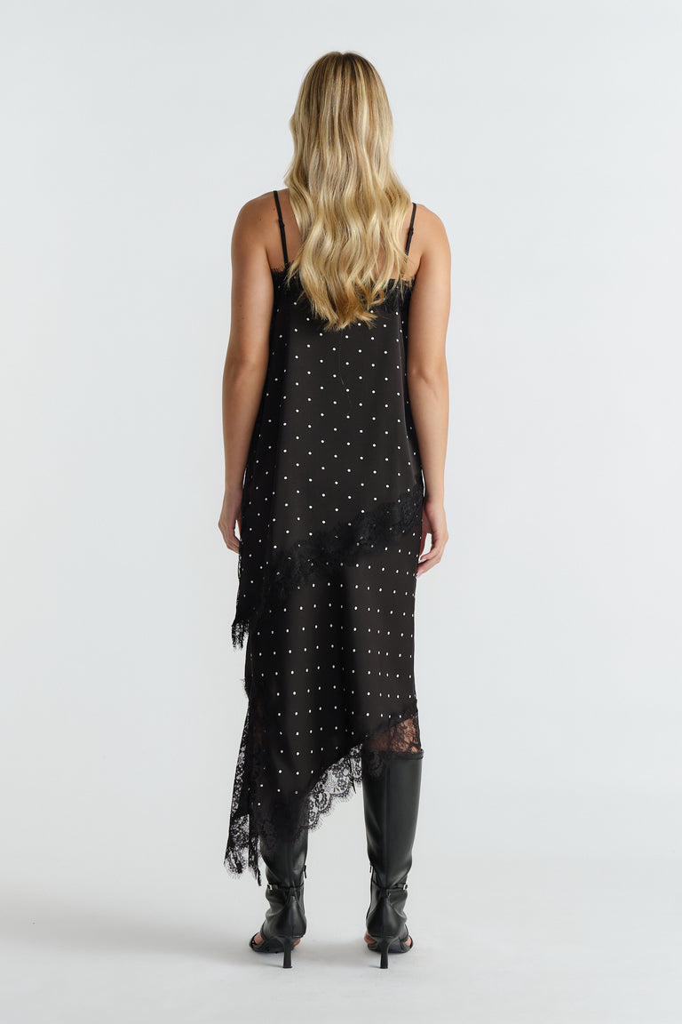 Woman wearing a black polka dot tunic with lace trim, thin straps, asymmetric hem, and black knee-high boots, back view
