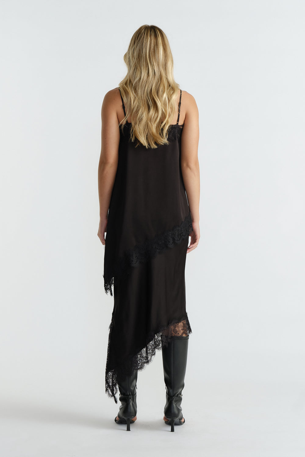 Back view of a woman wearing a black lace-trimmed tunic dress with an asymmetric hem and knee-high boots.