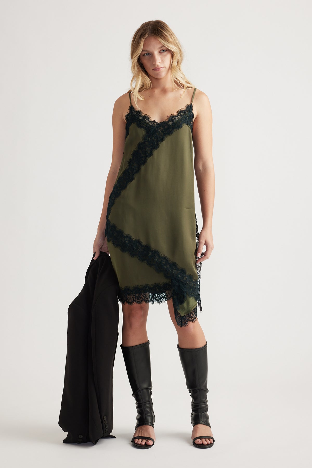 Woman wearing an olive slip dress with black lace trim, holding a black blazer and styled with black knee-high sandals