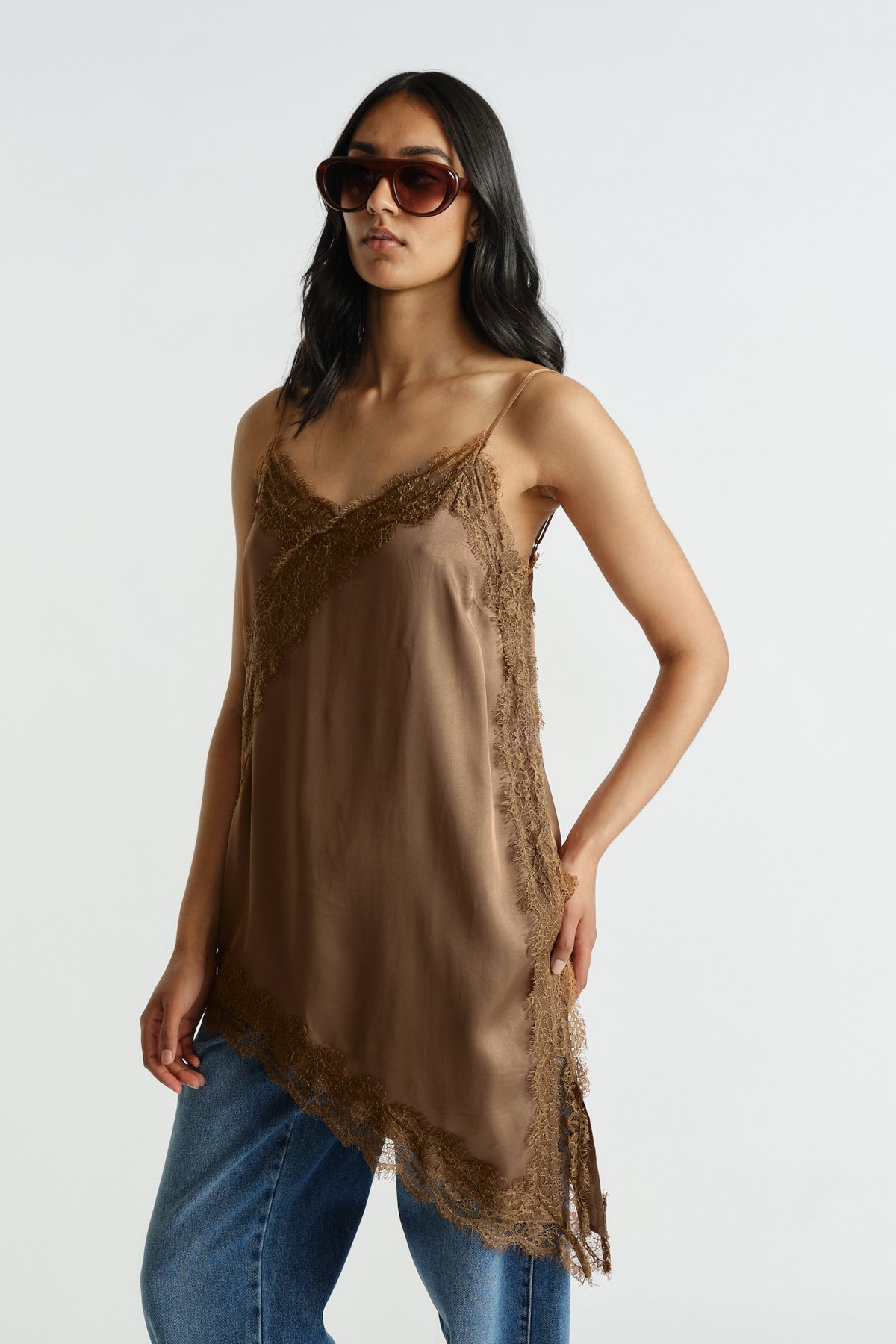 Woman wearing a brown lace-trimmed tunic with thin straps and asymmetric hem paired with blue denim jeans and sunglasses