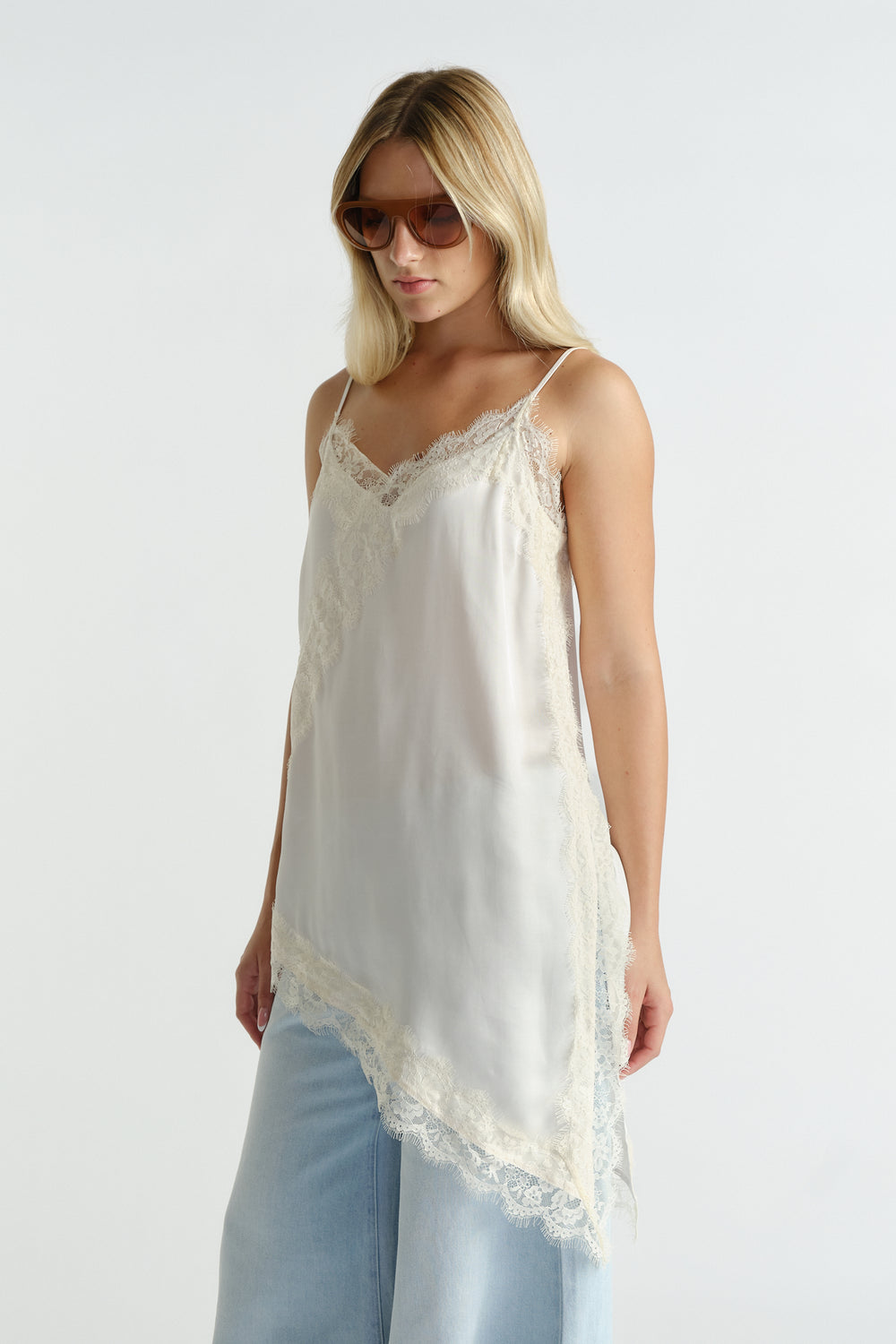 Woman wearing a cream lace-trimmed silk tunic layered over light blue wide-leg jeans and oversized brown sunglasses