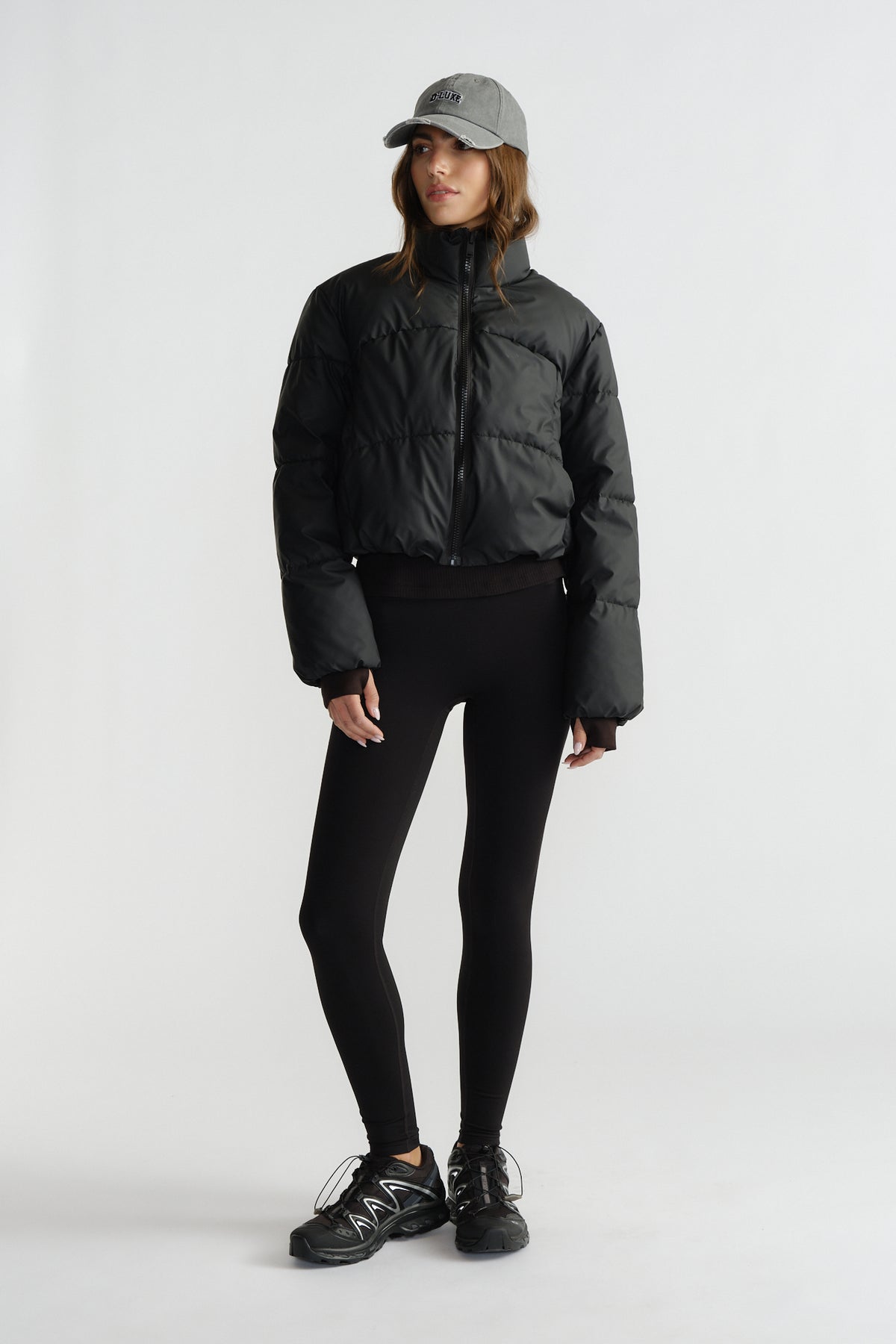 Woman wearing fitted black leggings paired with a cropped puffer jacket, sneakers, and a grey baseball cap for a casual look