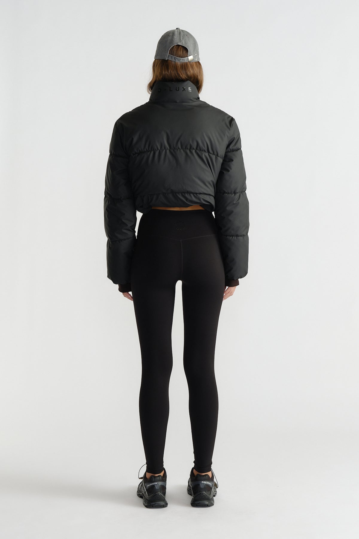 Rear view of woman wearing fitted black leggings paired with a cropped puffer jacket and a grey baseball cap