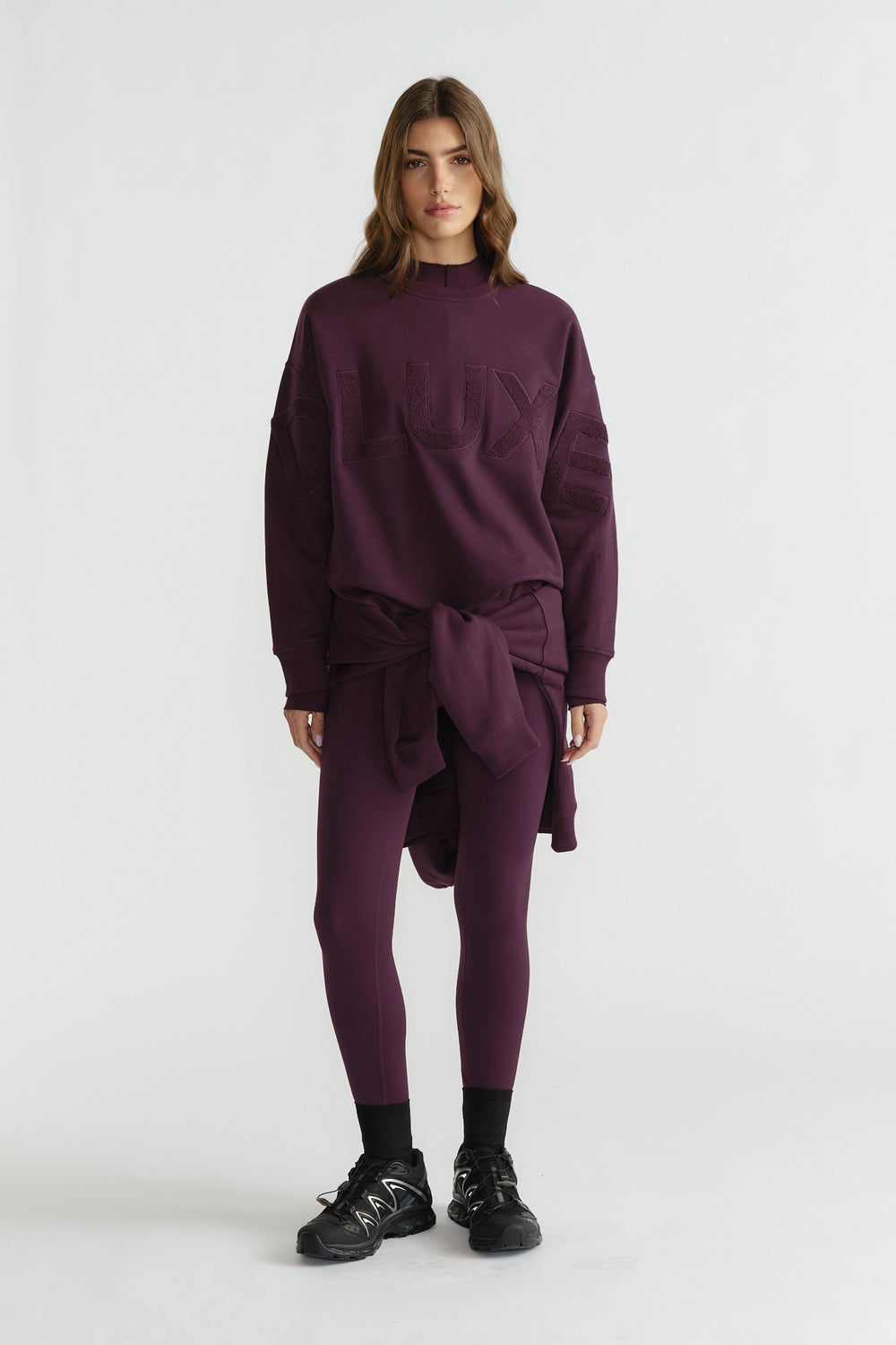 Woman wearing deep plum leggings and oversized matching sweatshirt tied at the waist, paired with black sneakers and socks