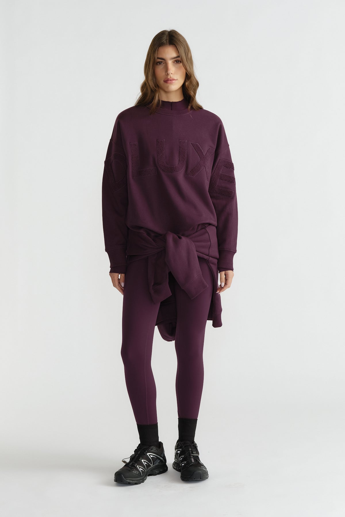 Woman wearing deep plum leggings and oversized matching sweatshirt tied at the waist, paired with black sneakers and socks
