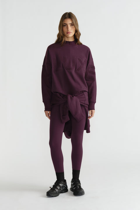 Woman wearing deep plum leggings and oversized matching sweatshirt tied at the waist, paired with black sneakers and socks