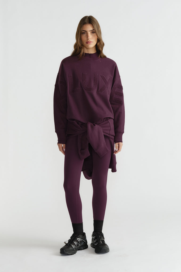 Woman wearing deep plum leggings and oversized matching sweatshirt tied at the waist, paired with black sneakers and socks