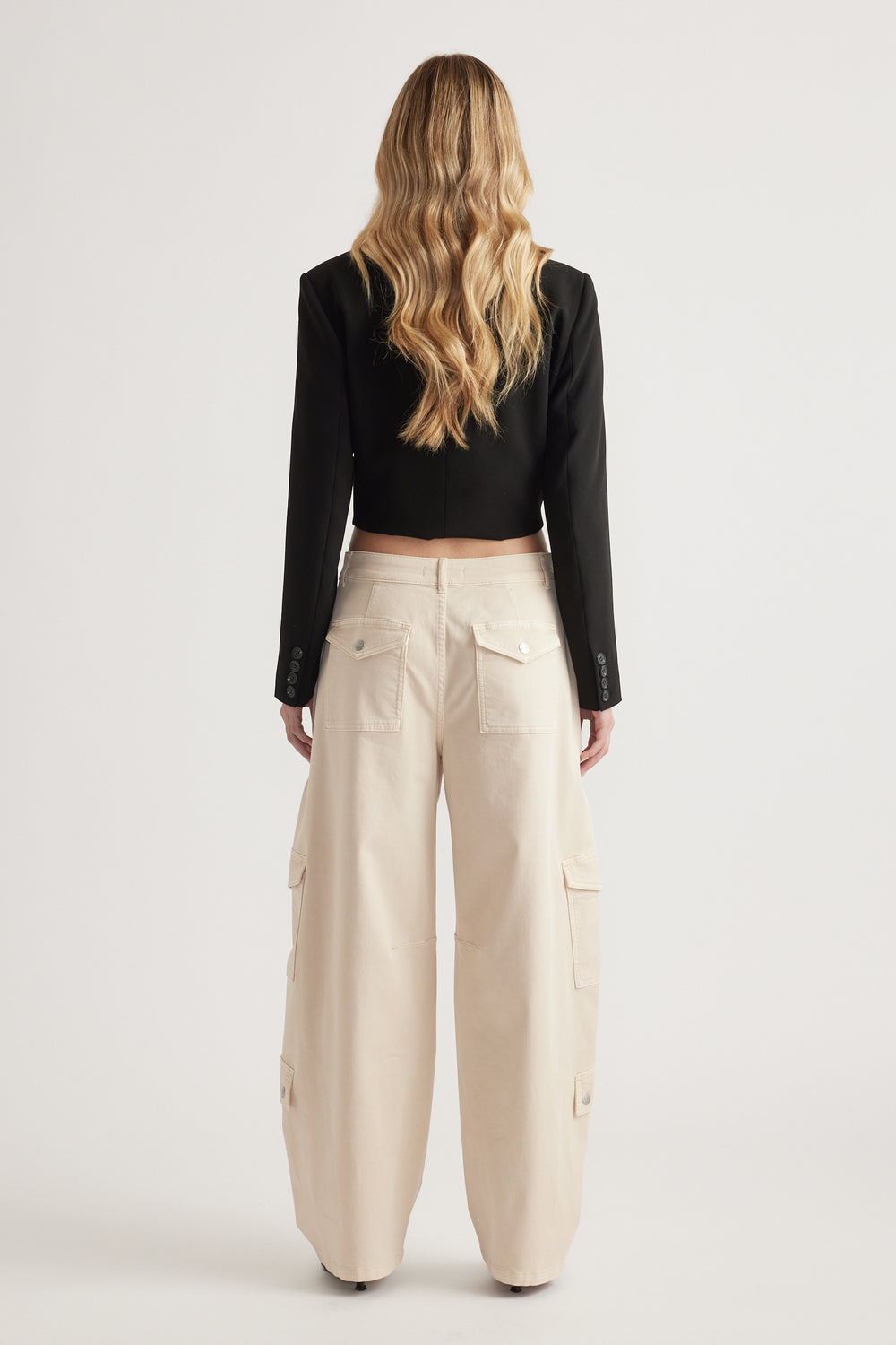 Back view of woman wearing cream barrel leg cargo pants with cargo pockets and a cropped black blazer