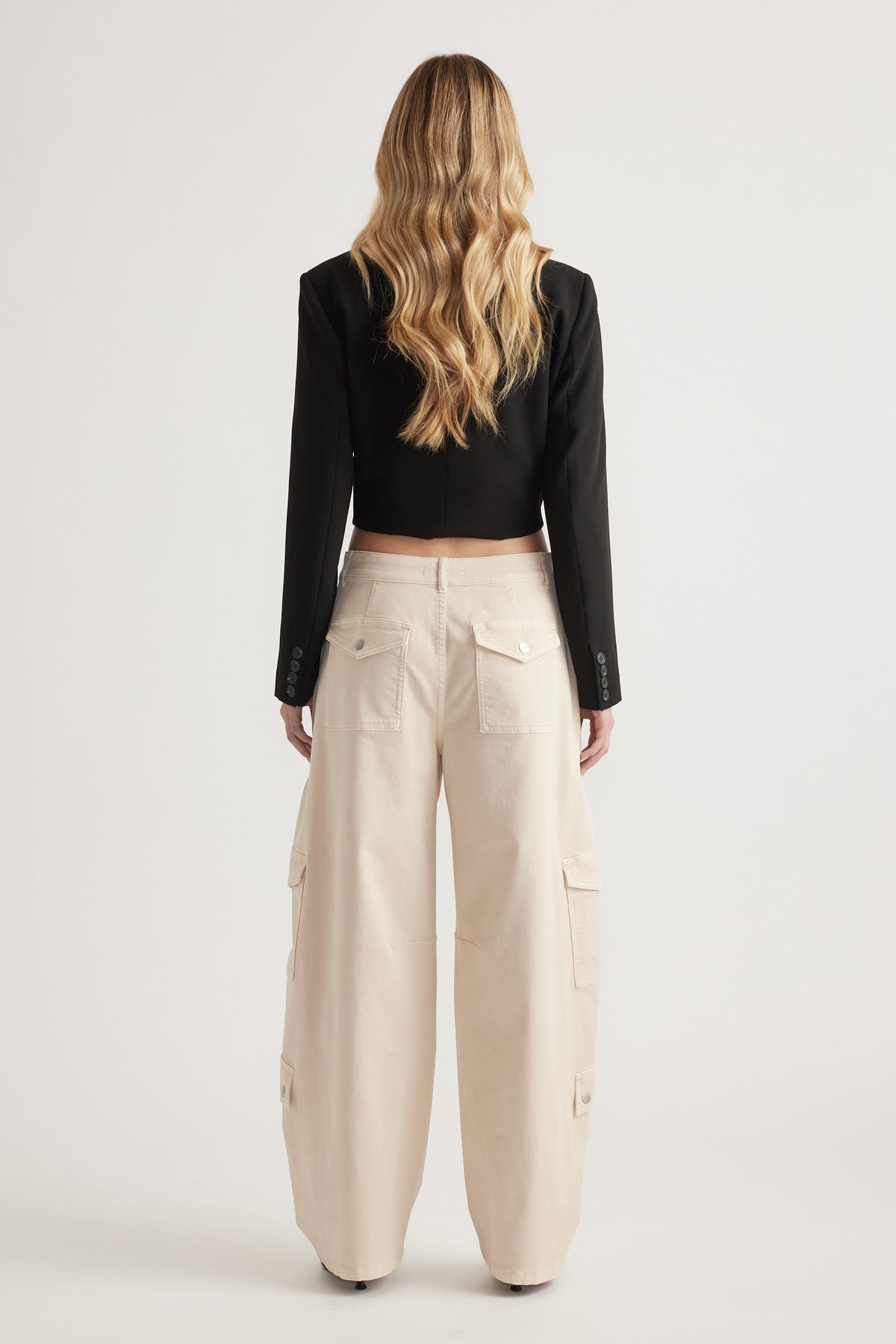 Back view of woman wearing cream barrel leg cargo pants with cargo pockets and a cropped black blazer