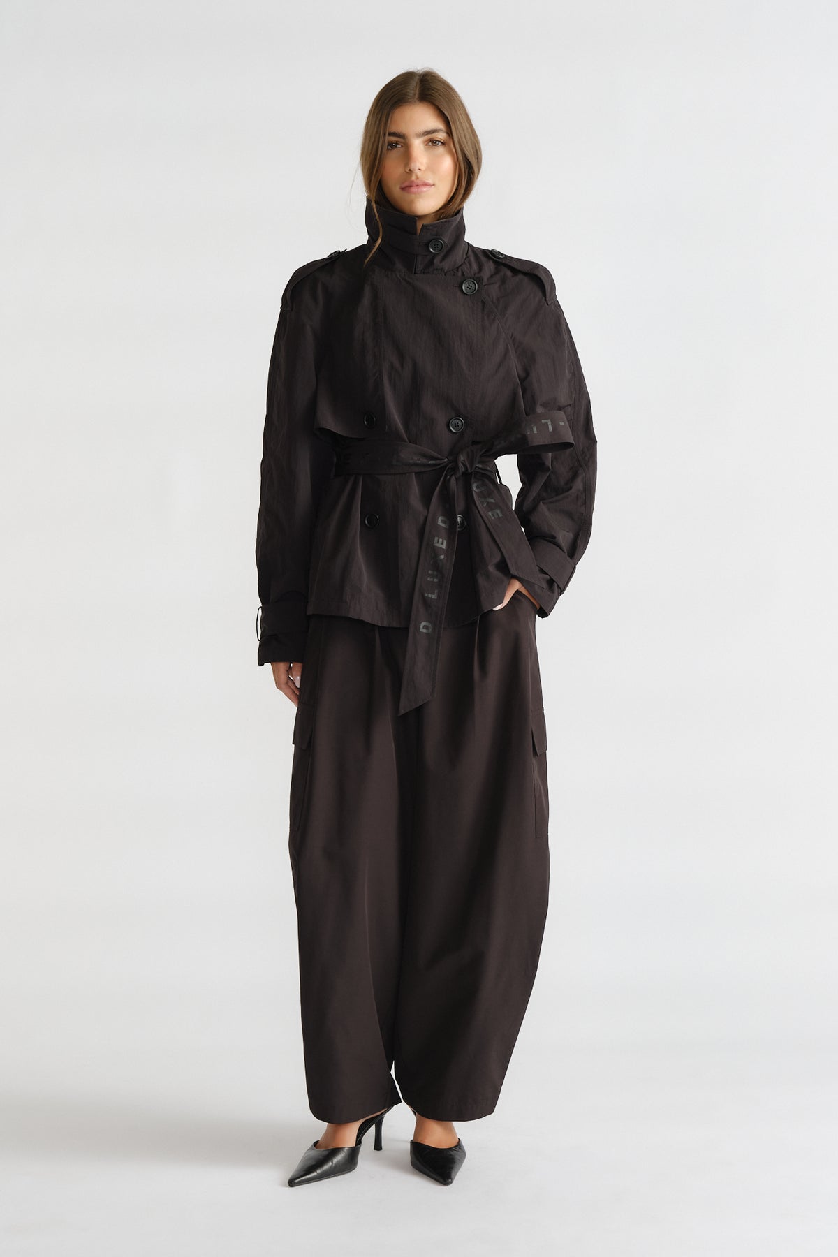 Woman wearing a black cropped trench with a waist tie, paired with wide-leg trousers and pointed black heels.