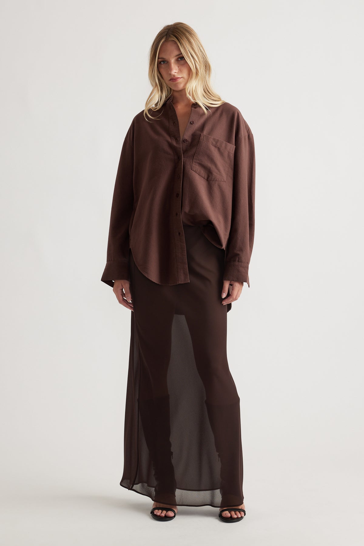 Woman wearing an oversized chocolate brown shirt with a chest pocket paired with a sheer long brown skirt and black sandals