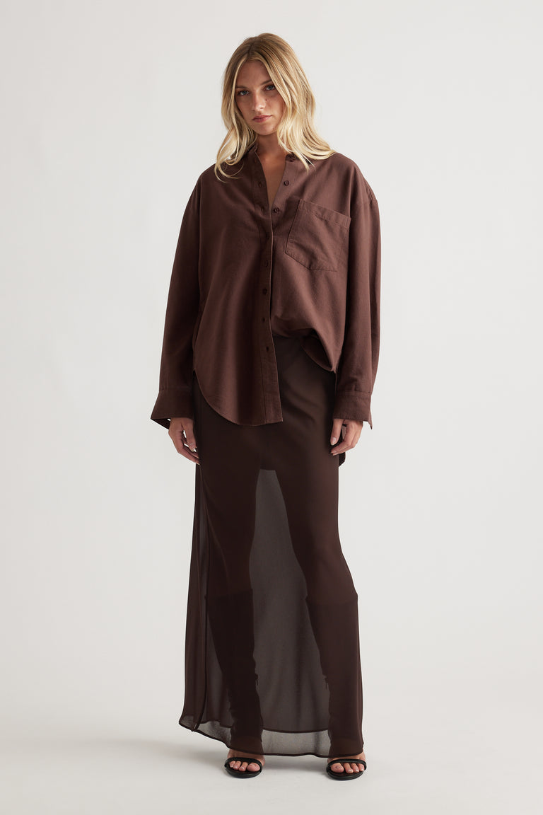 Woman wearing an oversized chocolate brown shirt with a chest pocket paired with a sheer long brown skirt and black sandals