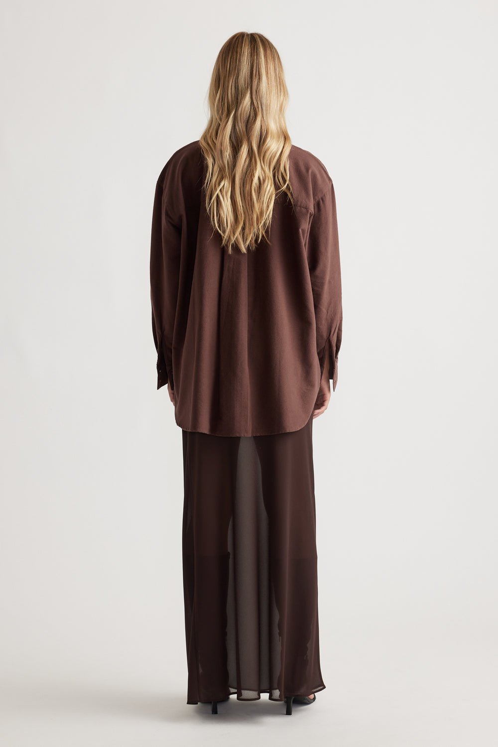 Woman seen from behind wearing a relaxed fit chocolate brown oversized shirt with long sleeves and a sheer flowing skirt