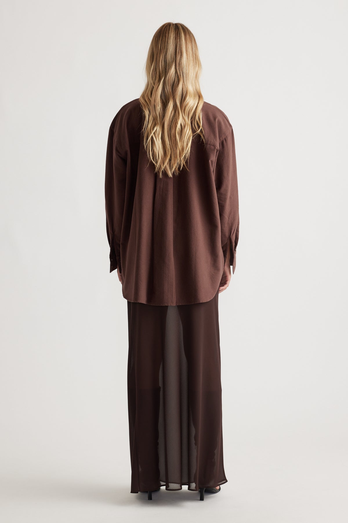 Woman seen from behind wearing a relaxed fit chocolate brown oversized shirt with long sleeves and a sheer flowing skirt