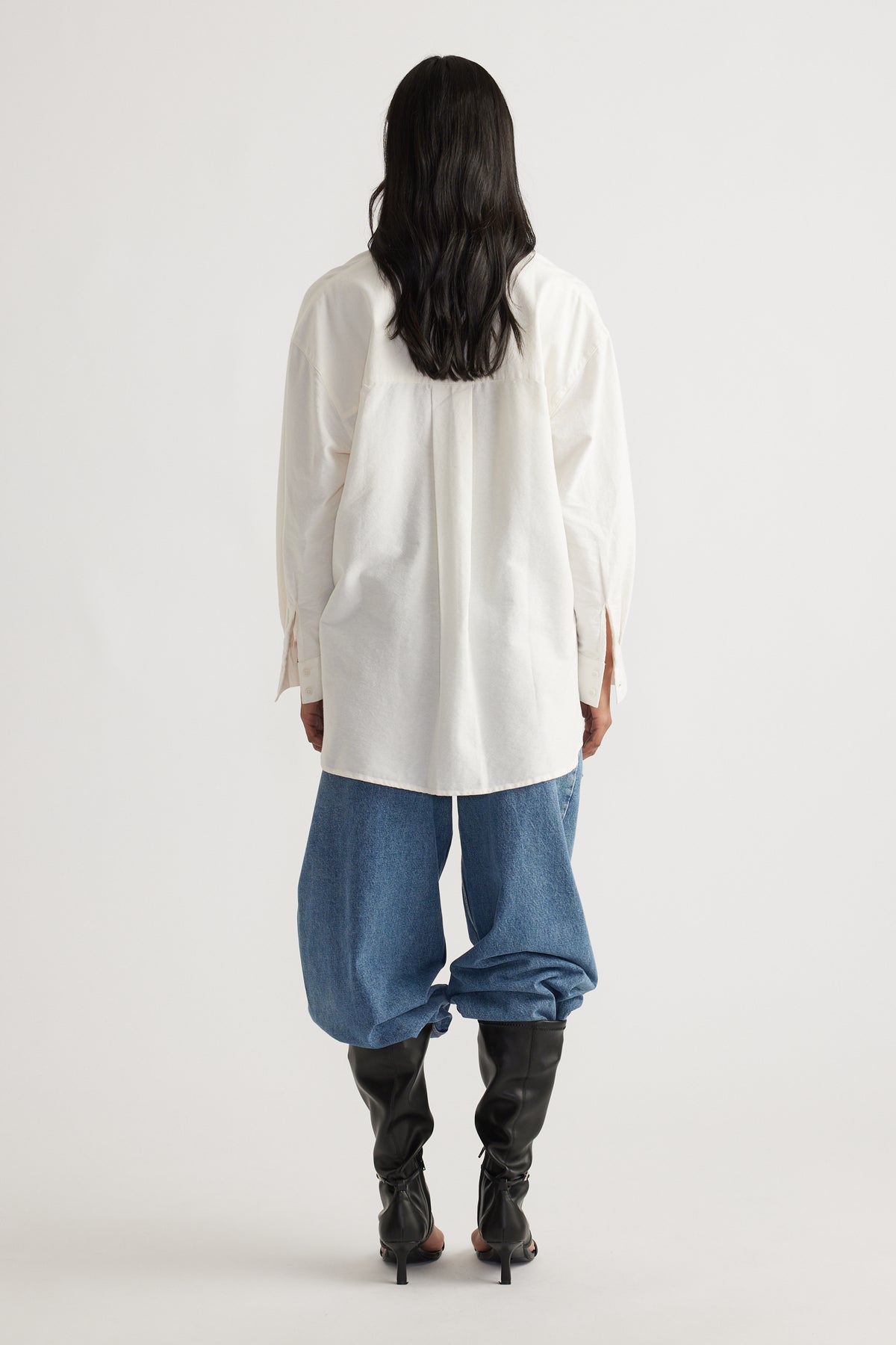 Back view of a relaxed warm white oversized shirt paired with loose blue denim jeans and black heeled boots.