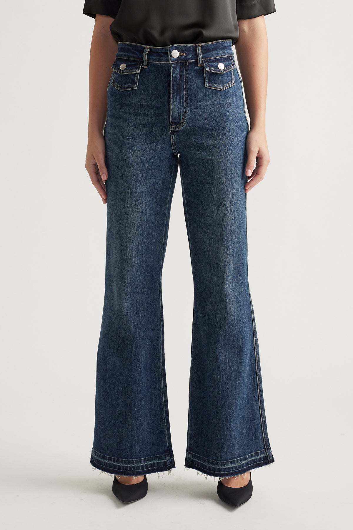 Woman wearing dark blue high-waisted flare jeans with front flap pockets and a black top paired with black pointed flats.