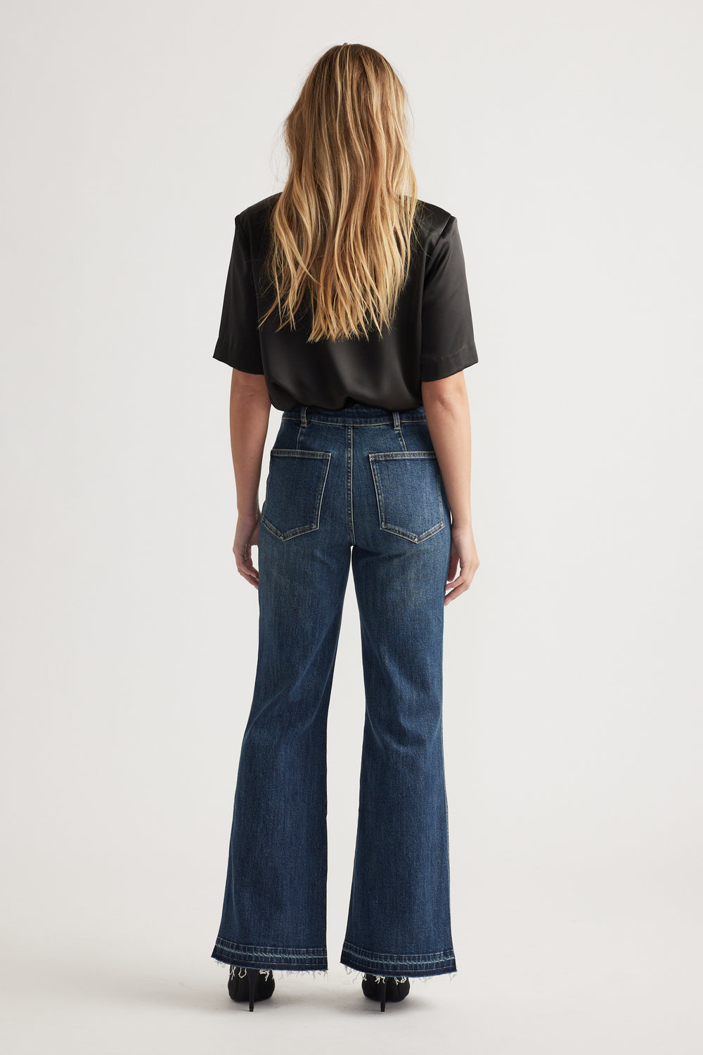 Woman with loose blonde hair wearing dark wash flare jeans and a black short-sleeve blouse, standing against a plain background.