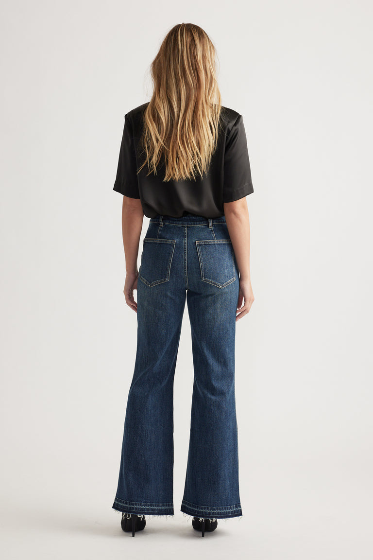 Woman with loose blonde hair wearing dark wash flare jeans and a black short-sleeve blouse, standing against a plain background.