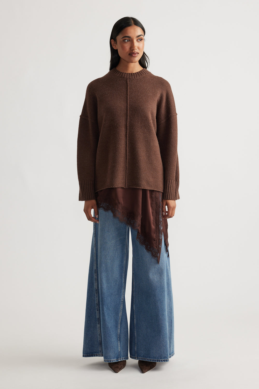 Woman wearing a coffee brown knit jumper with seam detail layered over a brown lace-trimmed top and wide-leg blue denim jeans