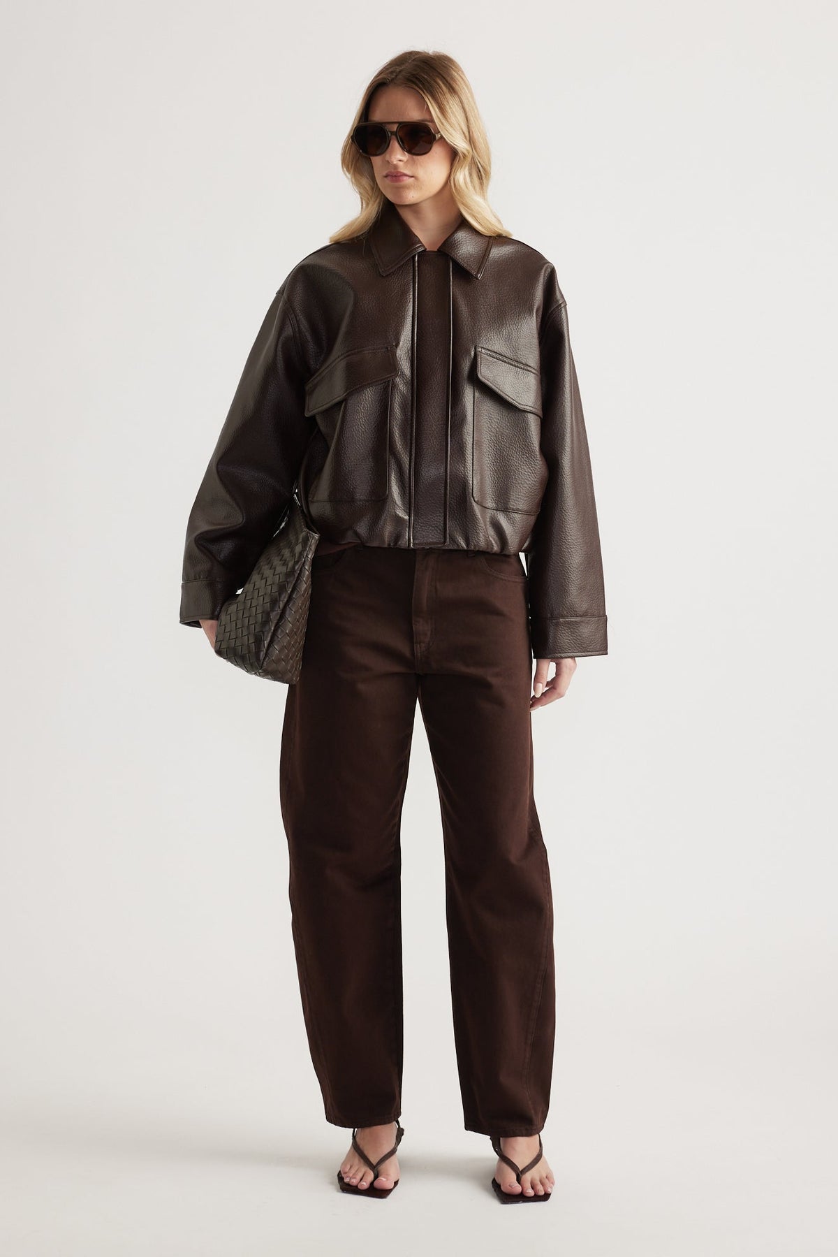 Woman wearing a dark brown faux leather bomber jacket with front pockets, paired with matching brown pants and sandals