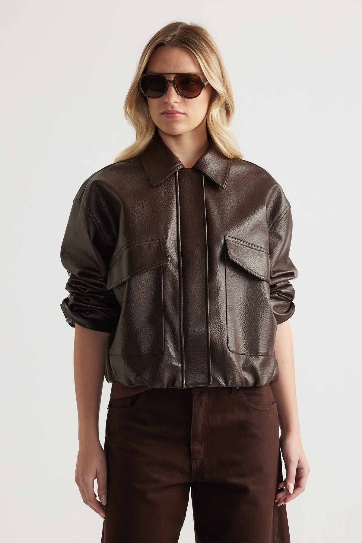 Woman wearing a dark brown faux leather bomber jacket with patch pockets and rolled-up sleeves paired with matching pants