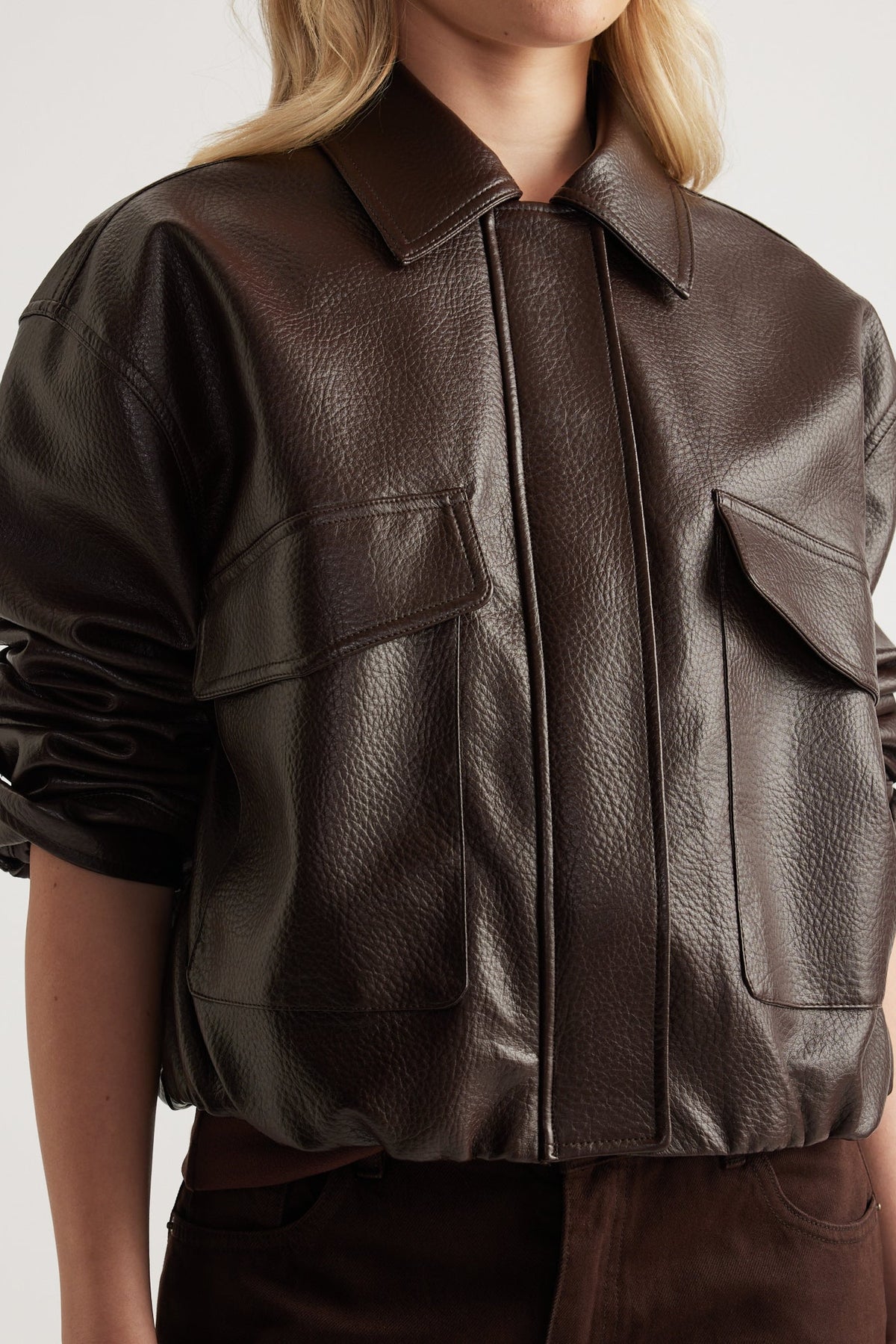 Close-up of dark brown faux leather jacket with collared neckline, flap front pockets, and ruched long sleeves worn by model