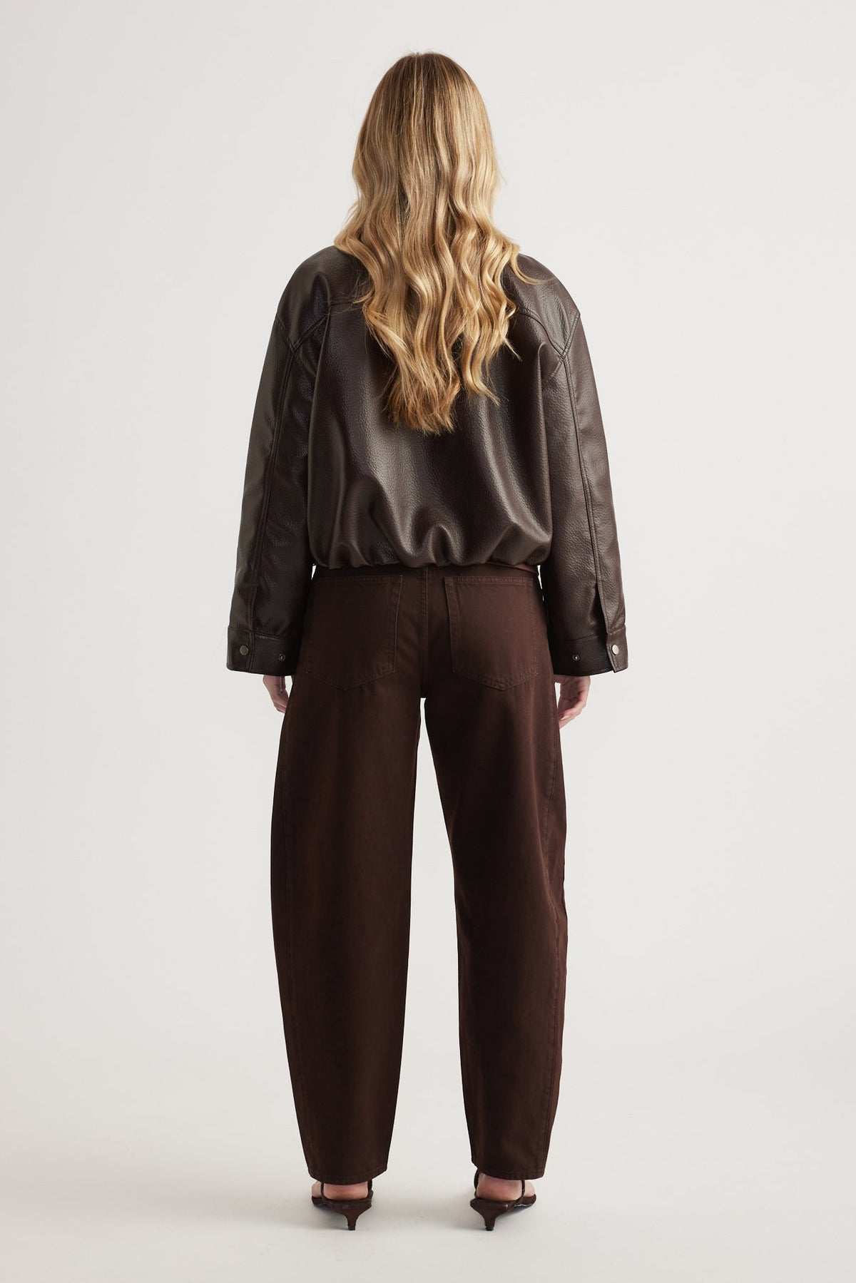 Woman seen from behind wearing a dark brown faux leather bomber jacket paired with loose dark brown trousers and heels