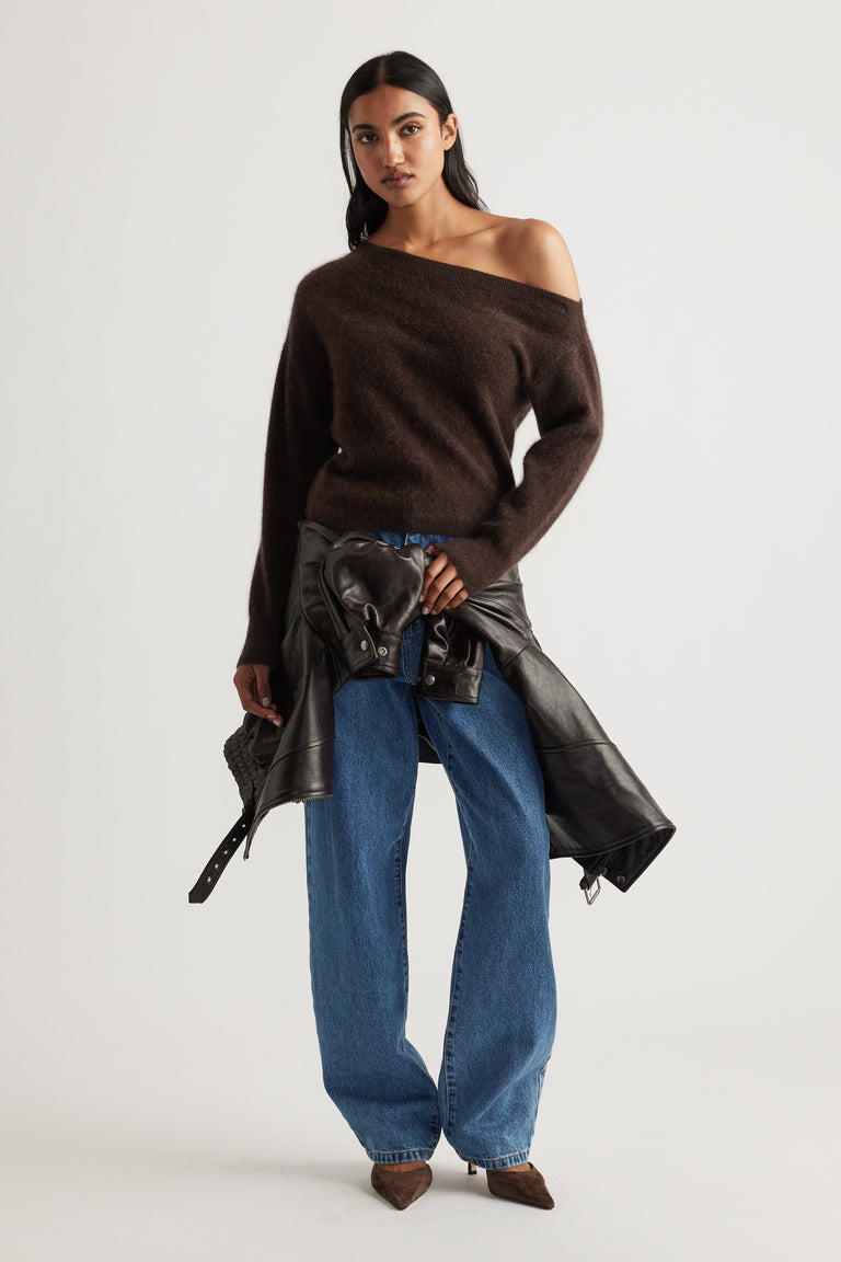 Woman wearing off-shoulder chocolate brown wool sweater paired with relaxed blue jeans and a tied black leather jacket.