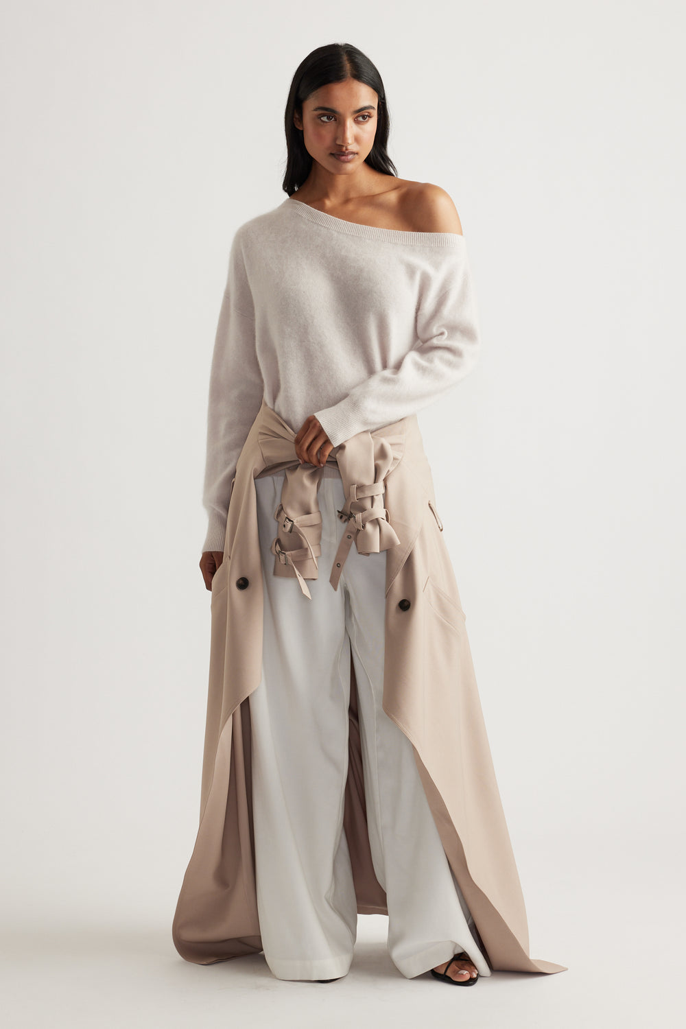 Woman wearing soft beige off-shoulder wool jumper paired with wide-leg white pants and a tied trench coat at the waist
