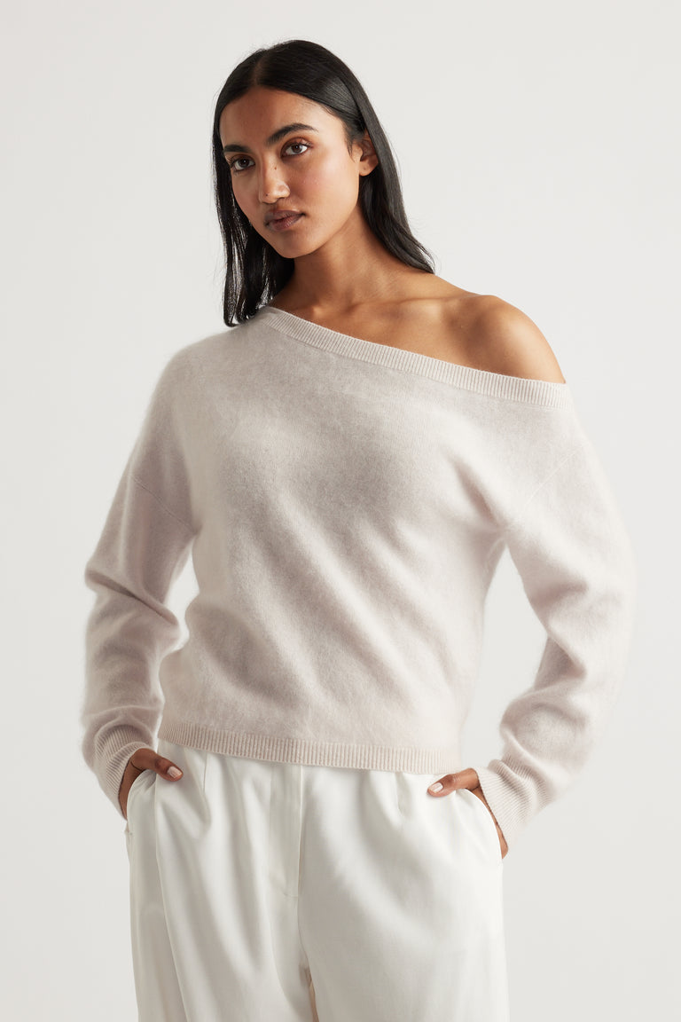 Woman wearing a soft neutral-toned wool jumper with a relaxed fit and one-shoulder neckline paired with white trousers