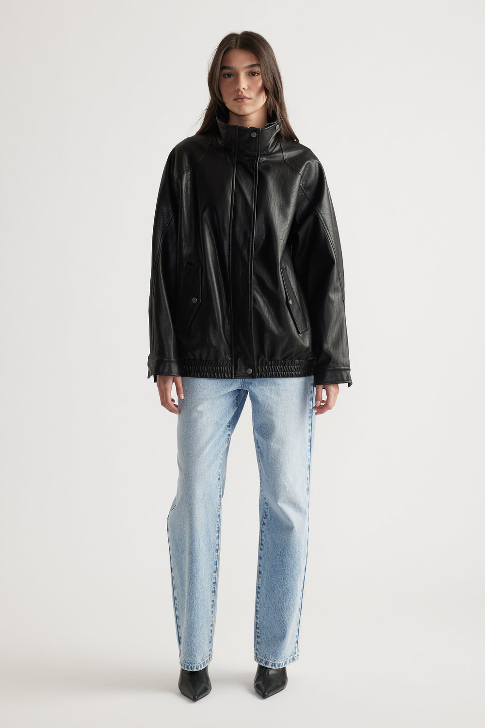 Woman wearing a black oversized faux leather jacket paired with light blue straight leg jeans and black pointed boots.