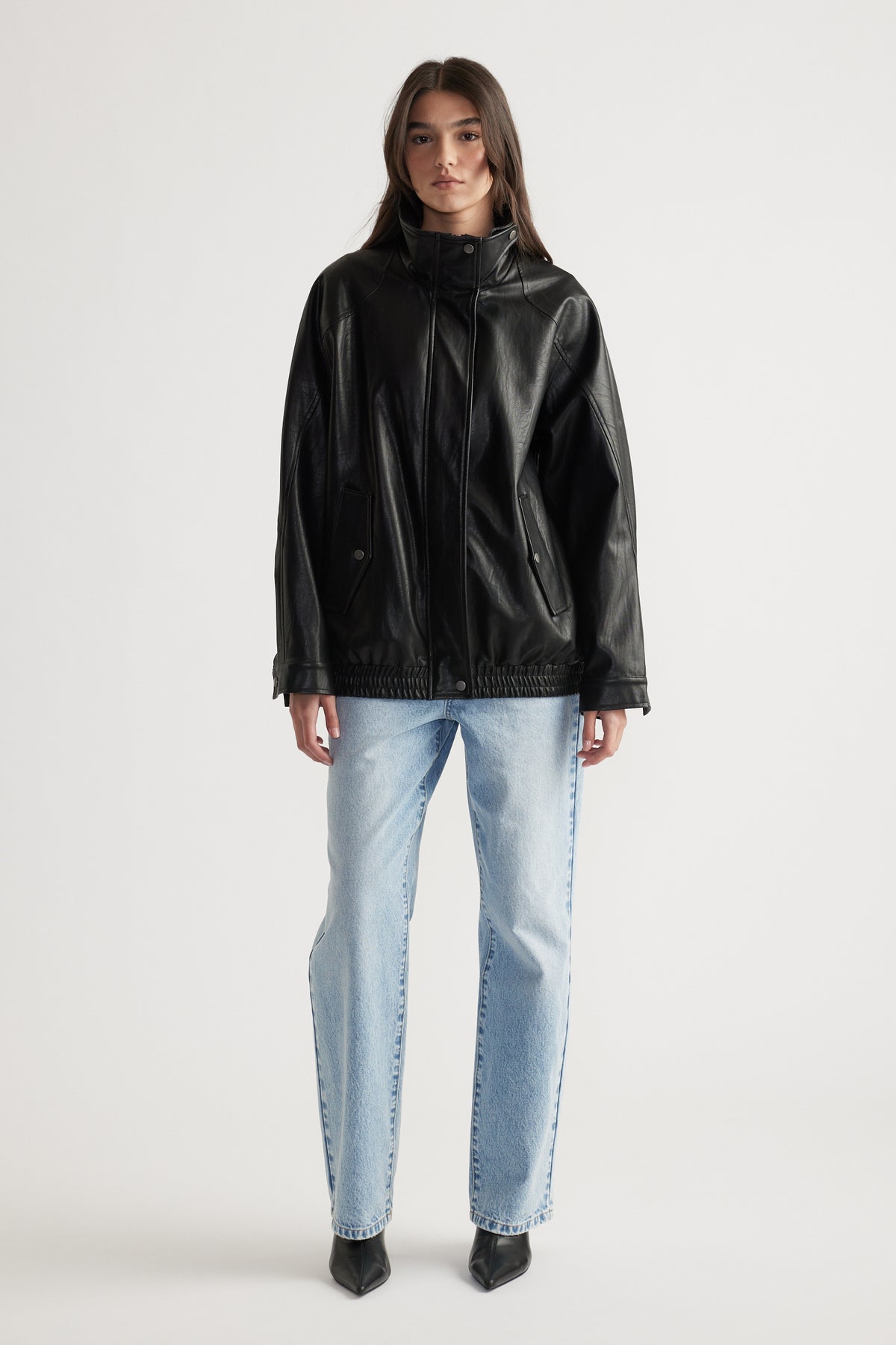 Woman wearing a black oversized faux leather jacket paired with light blue straight leg jeans and black pointed boots.
