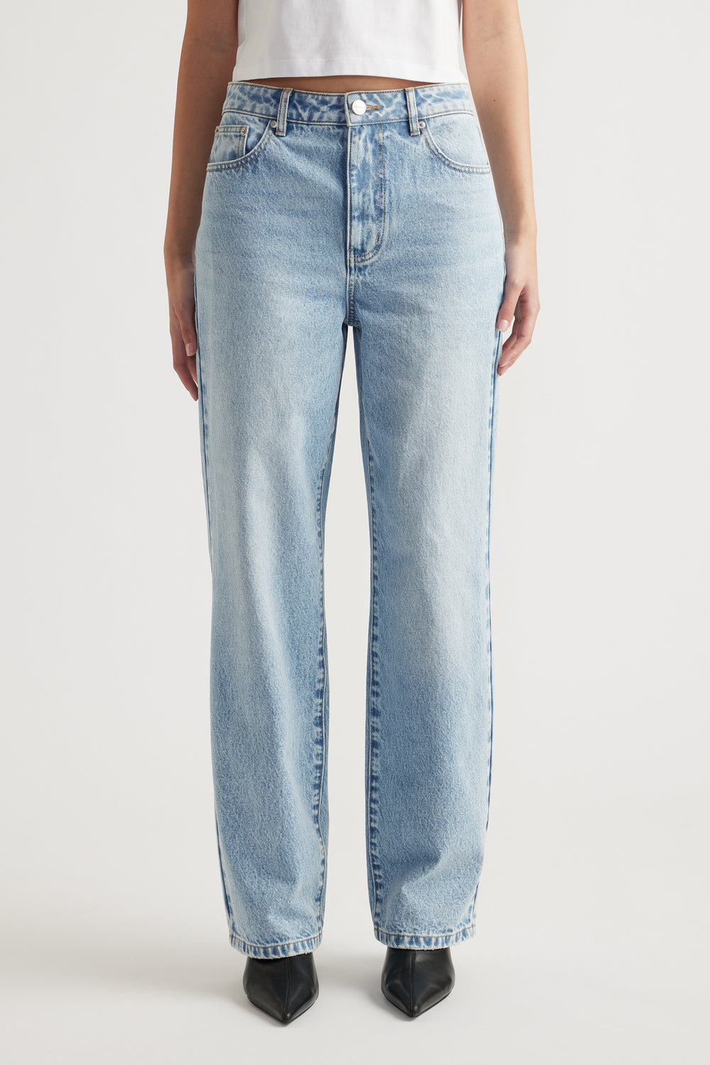 Woman wearing light blue straight leg jeans paired with black pointed boots and a white cropped top against a plain background.