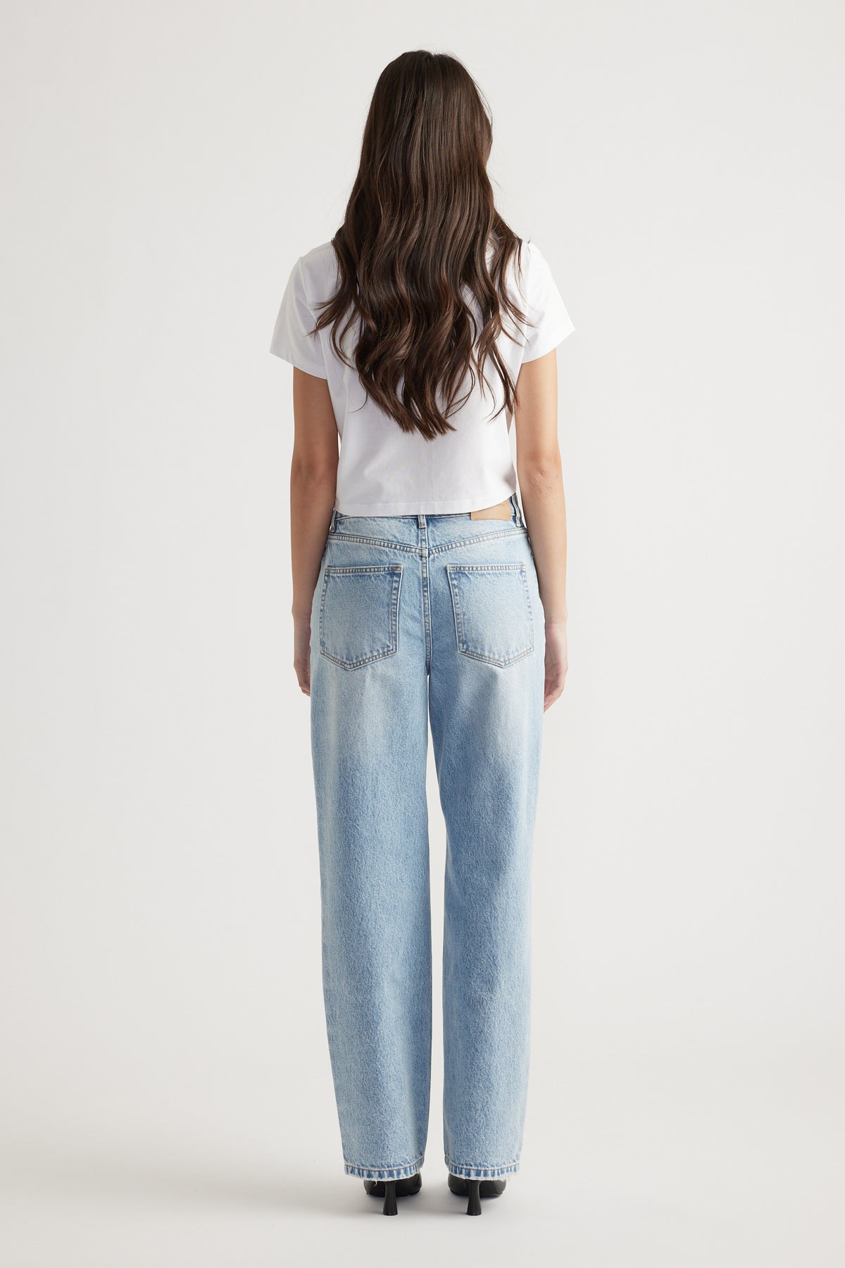 Woman with long wavy hair wearing light blue straight leg jeans and a white cropped T-shirt standing with back to camera.