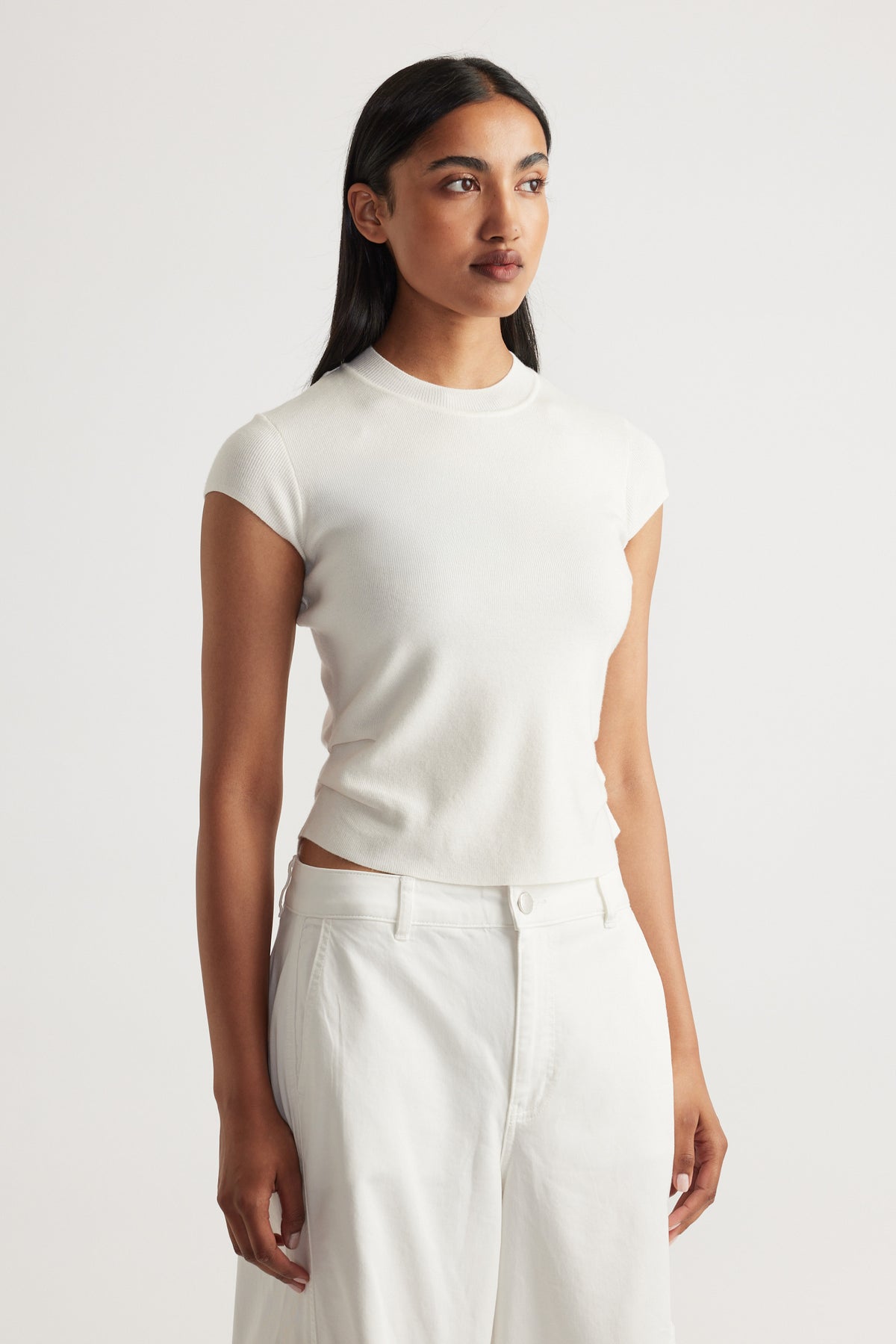 Woman wearing an ivory Lily Pleat Detail Knit Tee with cap sleeves styled with white pants by DECJUBA.