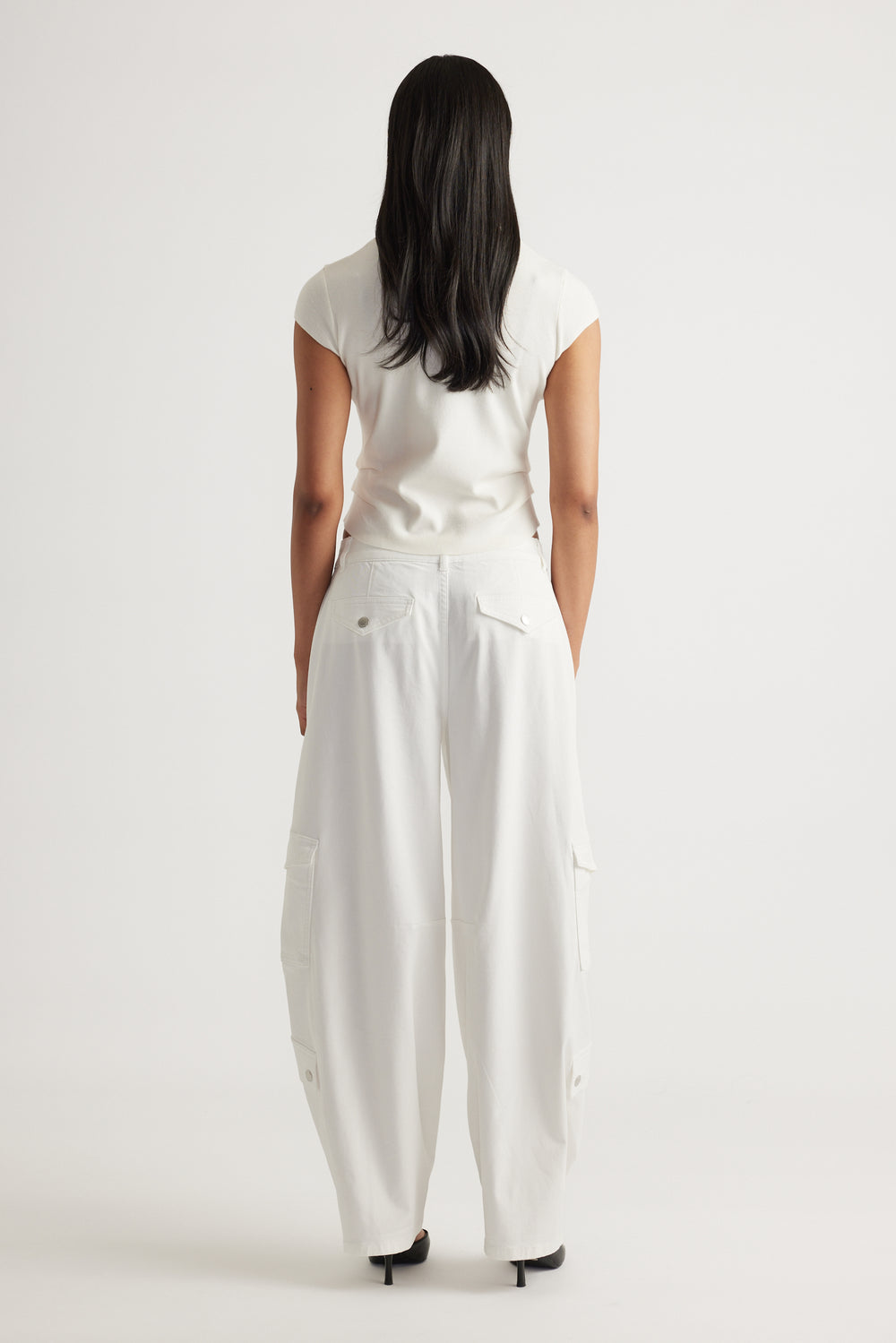 Rear view of a woman wearing the Lily Pleat Detail Knit Tee in soft winter white paired with wide-leg white pants by DECJUBA